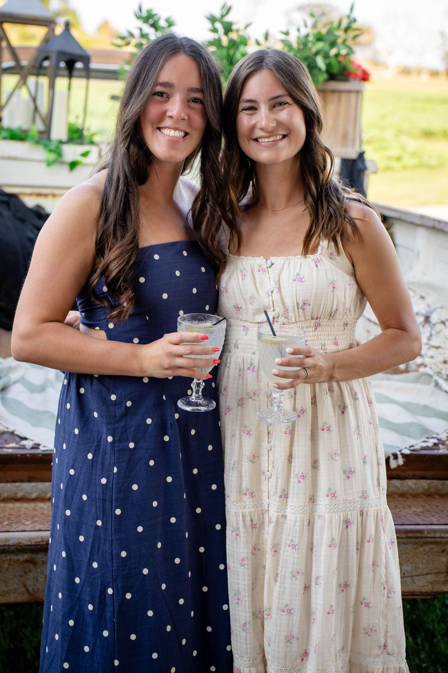 Two young women in summer maxi dresses smile while holding cocktails at an outdoor event.