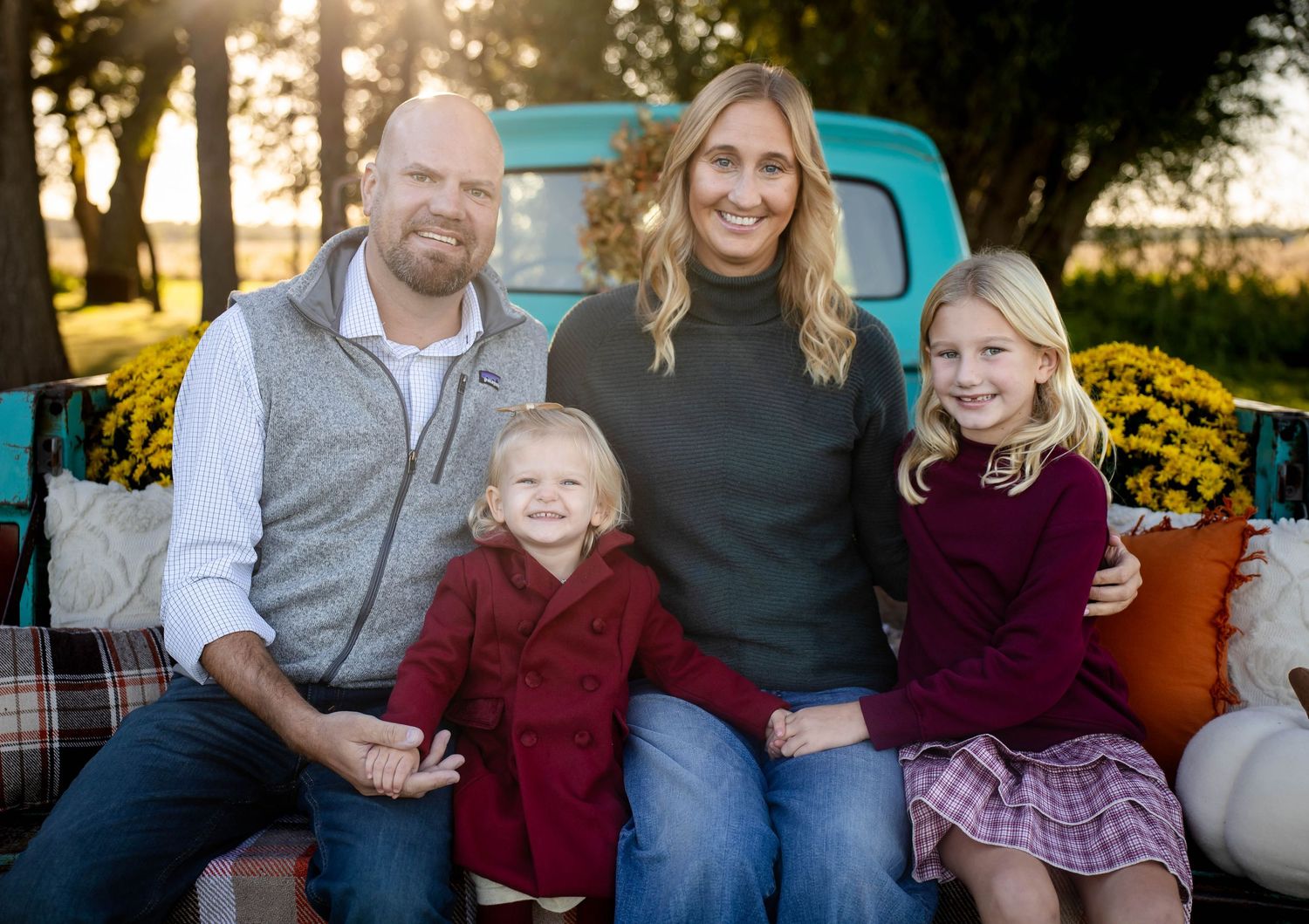 A smiling family of four sits together outdoors at sunset in front of a vintage teal truck, with yellow mums and a plaid blanket nearby.