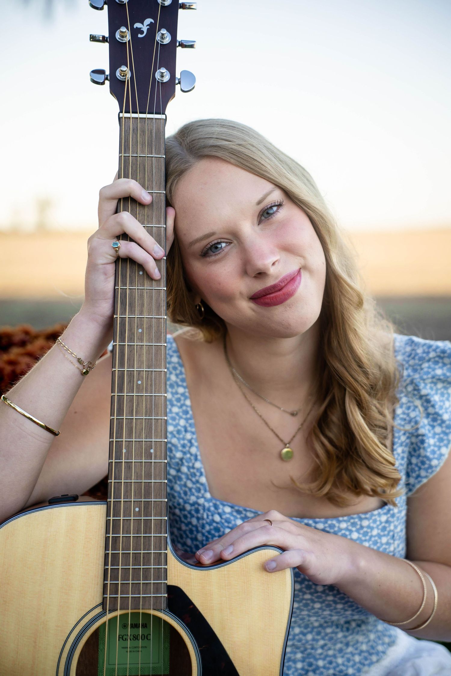 Smiling young woman with long blonde hair in a blue floral top rests her head against an acoustic guitar neck outdoors.