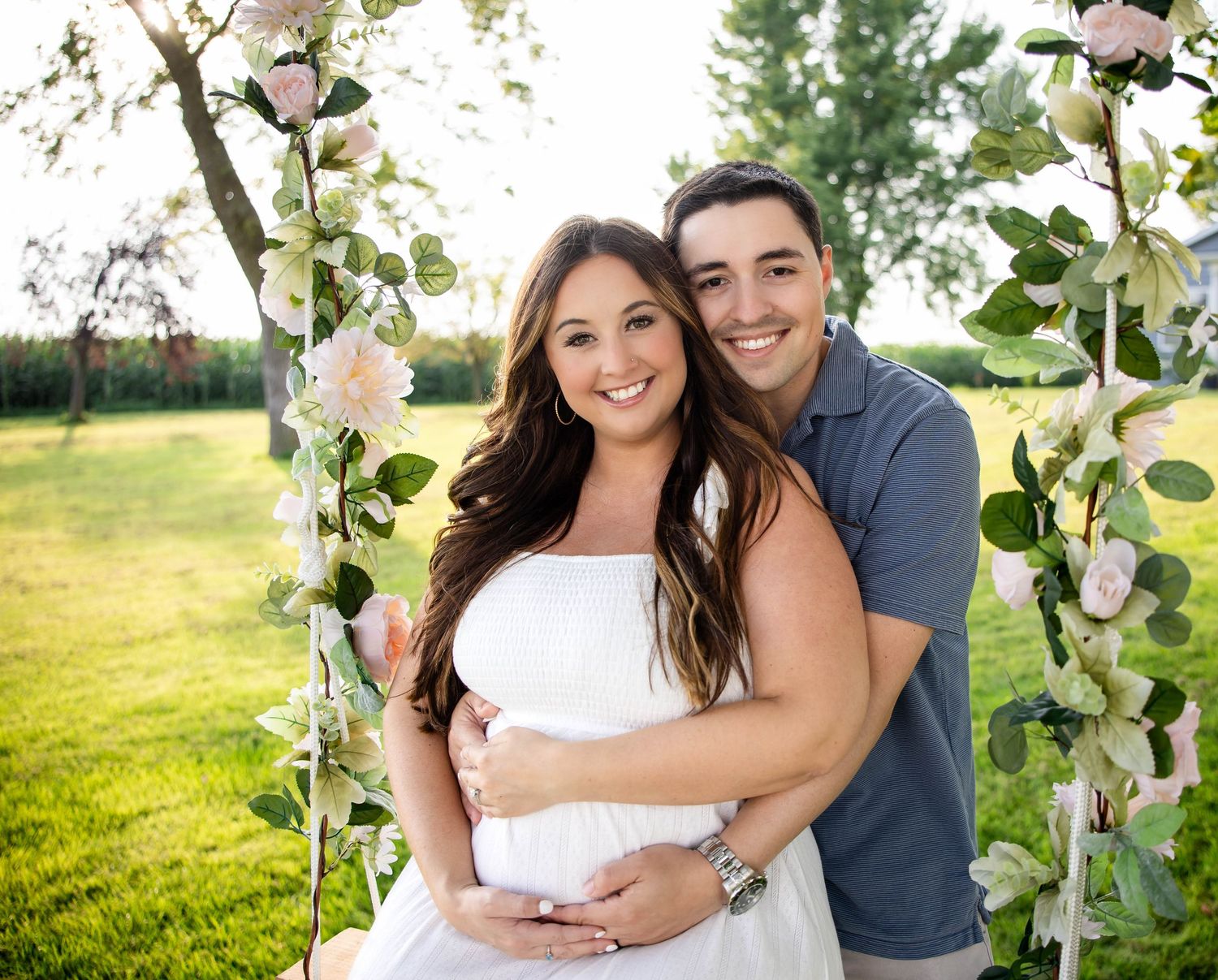 Smiling expecting couple embracing under a floral arch with pink roses in a garden setting.