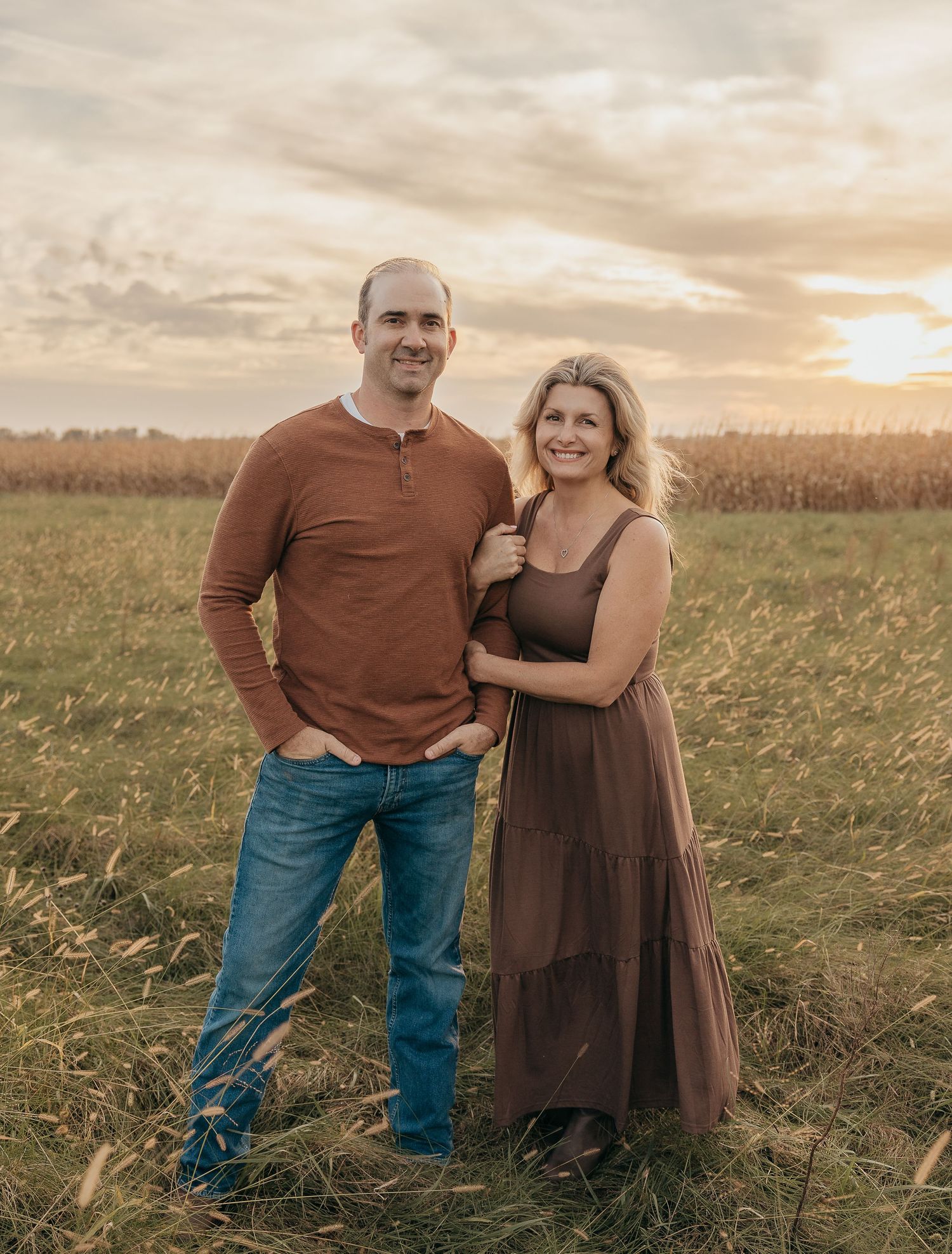 Couple posing together at sunset in a golden field, man in rust henley and woman in brown maxi dress.