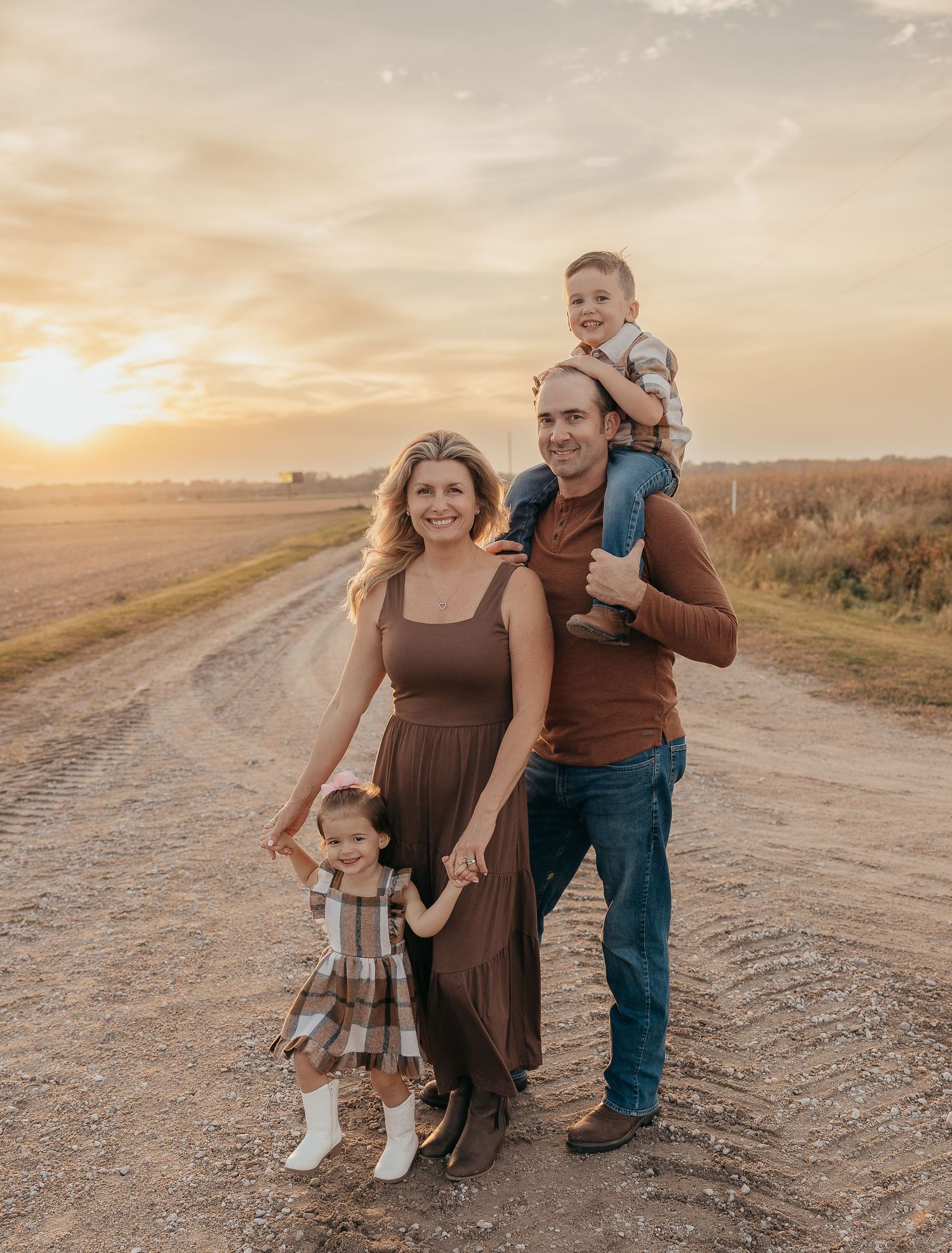 Family of four posing on a rural dirt road at golden hour sunset, mother in brown dress, father with son on shoulders.