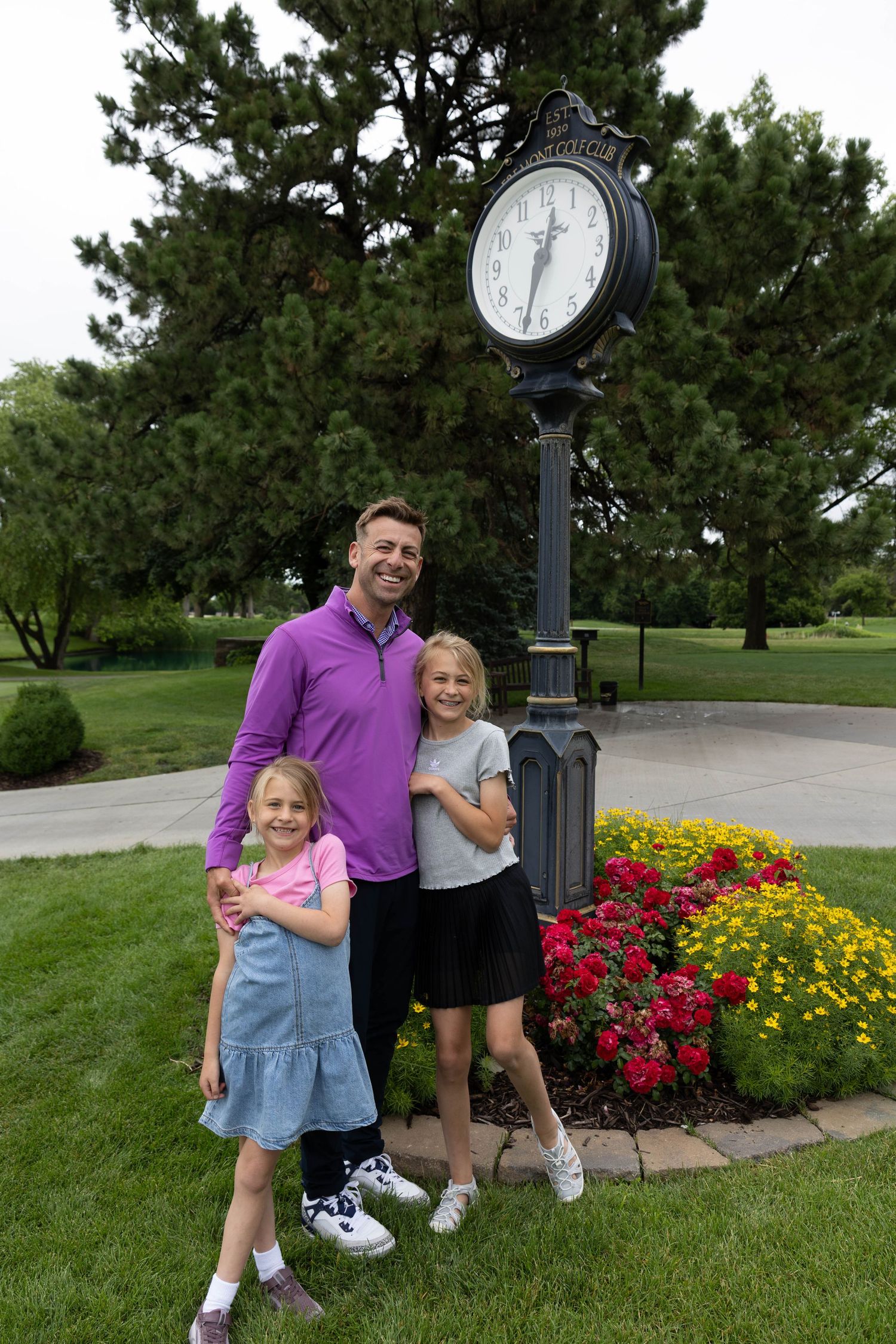 Man in purple polo shirt poses with two young girls beside decorative clock tower in park.