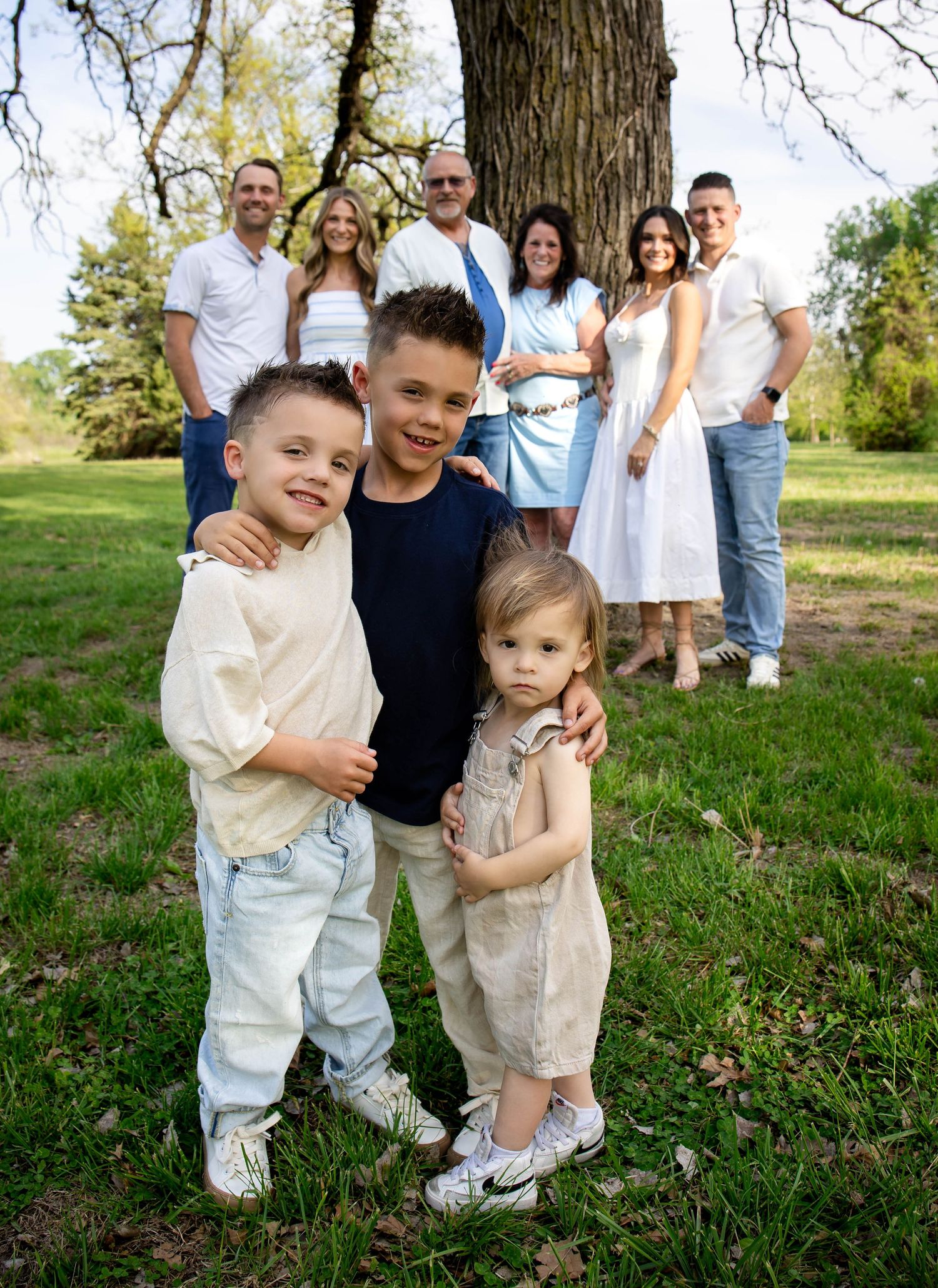 Extended family portrait with three young children in foreground and adults posed by a large tree.