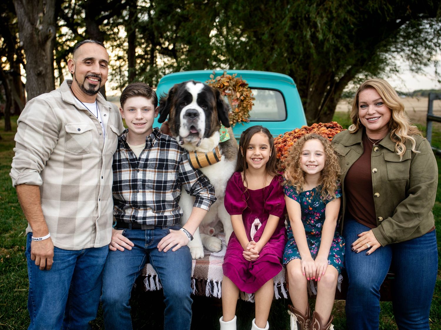 Family of five with two adults and three children posing outdoors beside a large St. Bernard dog and a vintage teal truck.