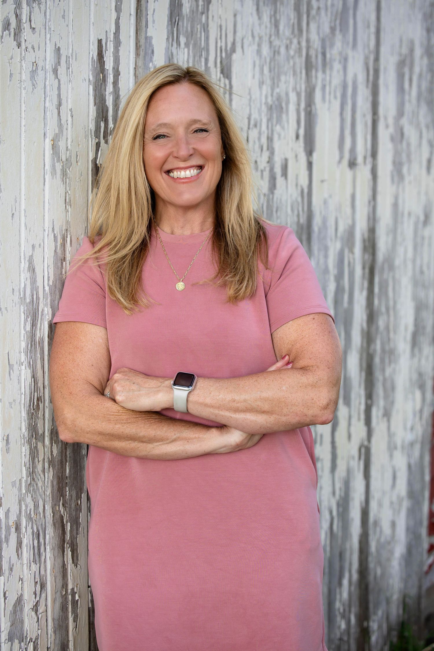 Smiling blonde woman in pink dress standing with arms crossed against a rustic white wooden wall.