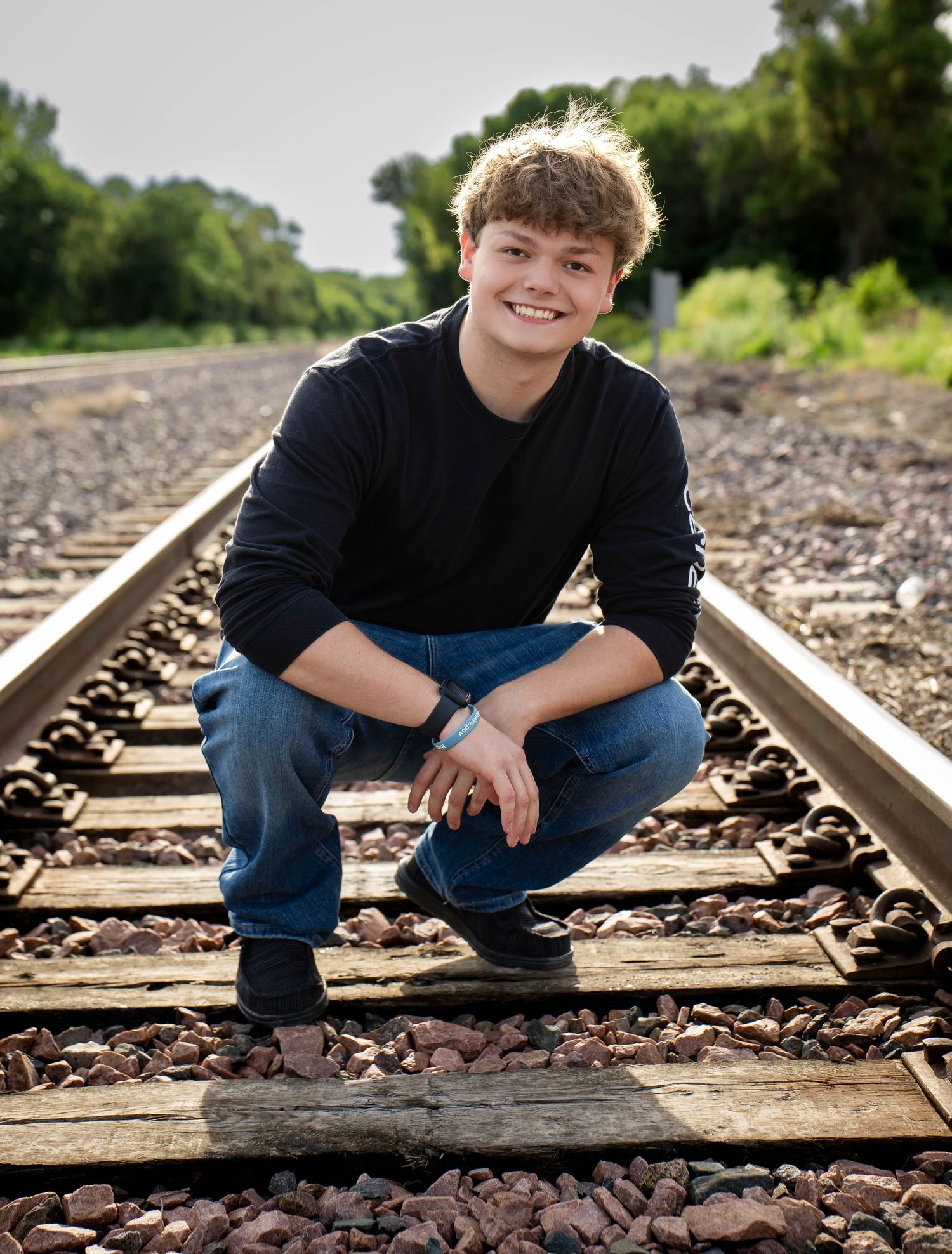 Smiling teenage boy crouching on railroad tracks surrounded by gravel, senior portrait photo.