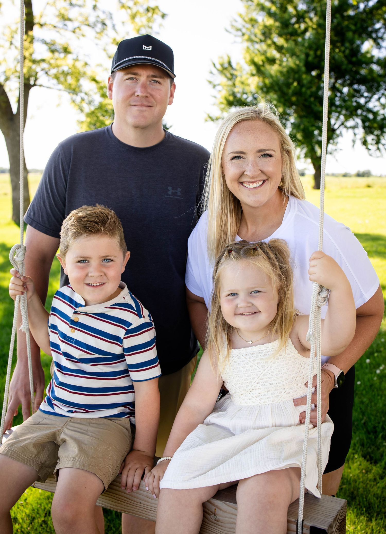 Family of four smiling together on wooden swing in lush green summer outdoor portrait session.