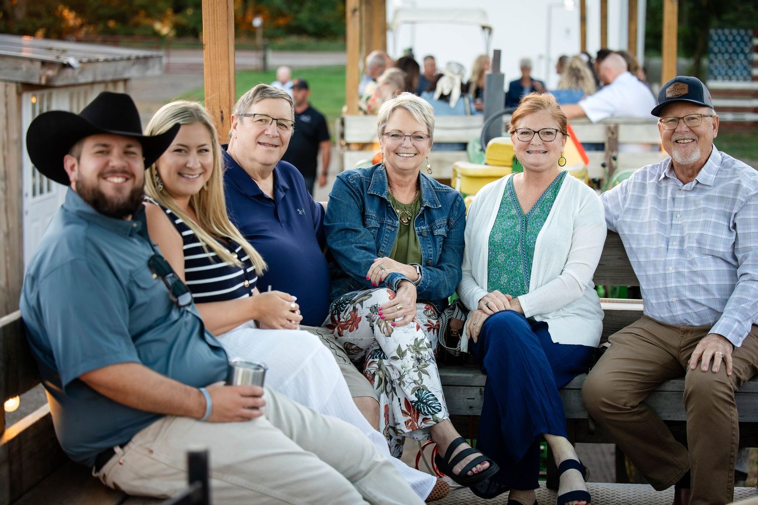 Group of six adults smiling together on a bench at an outdoor evening event with warm ambient lighting.