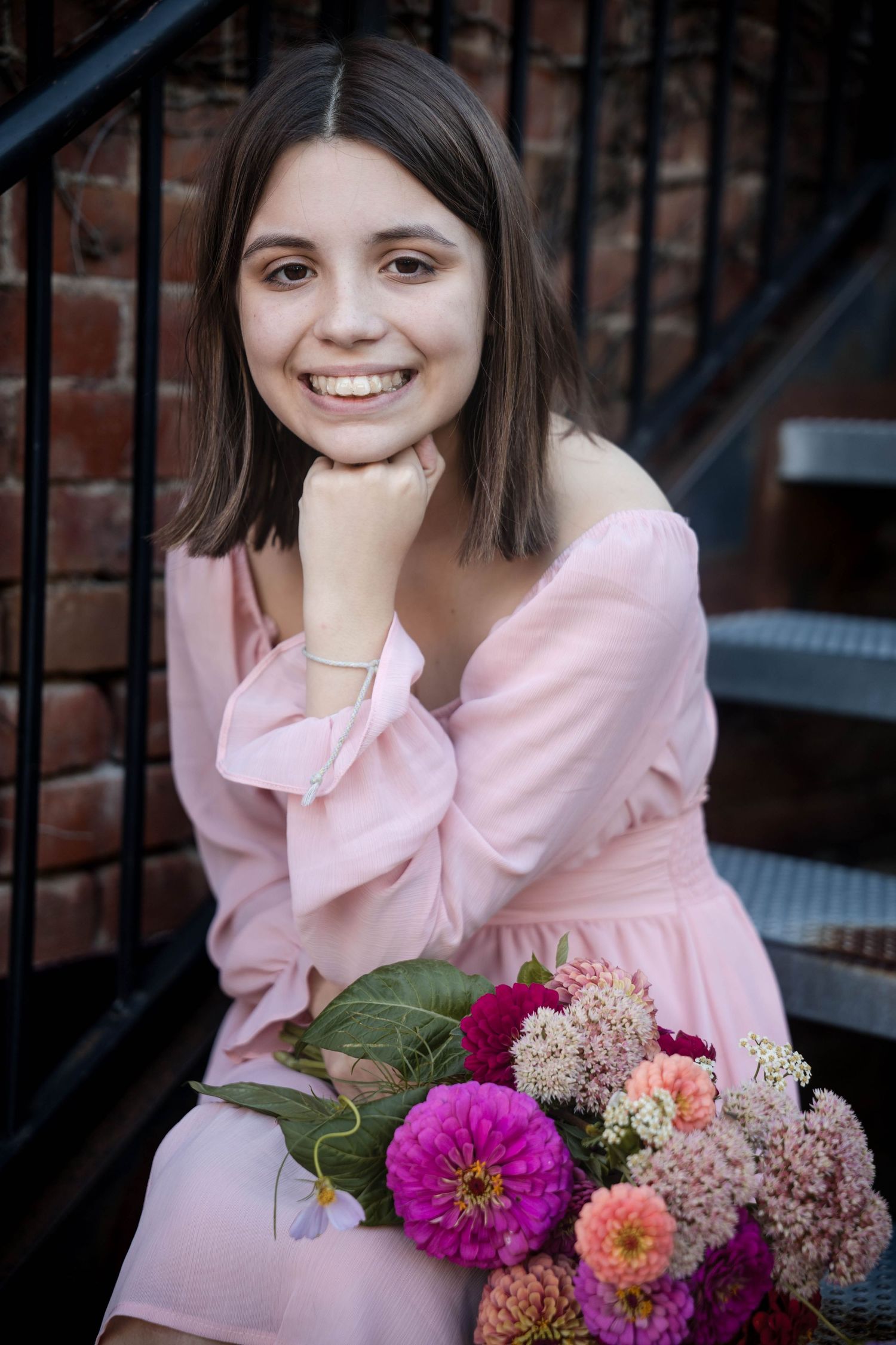Smiling young woman in a pink off-shoulder dress sits on outdoor steps near a brick wall holding a bouquet of pink and magenta flowers.