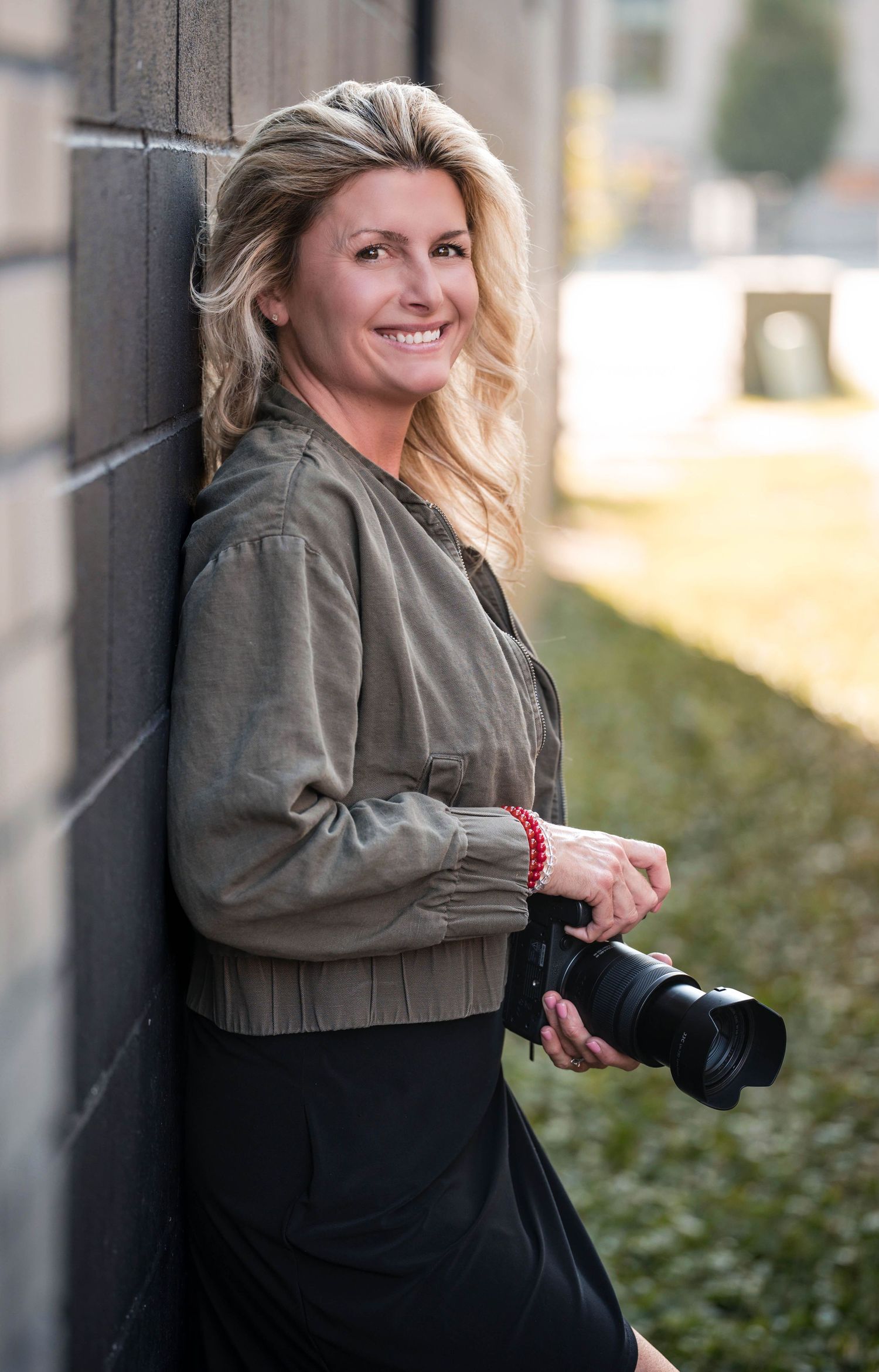Smiling blonde female photographer leaning against brick wall holding DSLR camera outdoors.