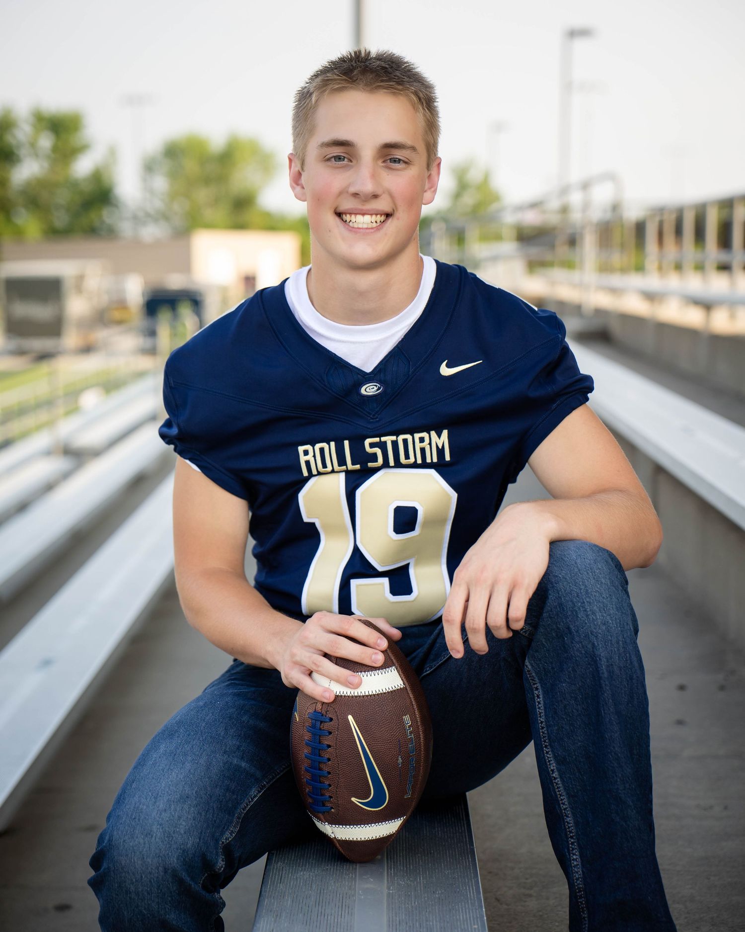 Smiling teen in Roll Storm football jersey number 19 holding football in stadium bleachers.