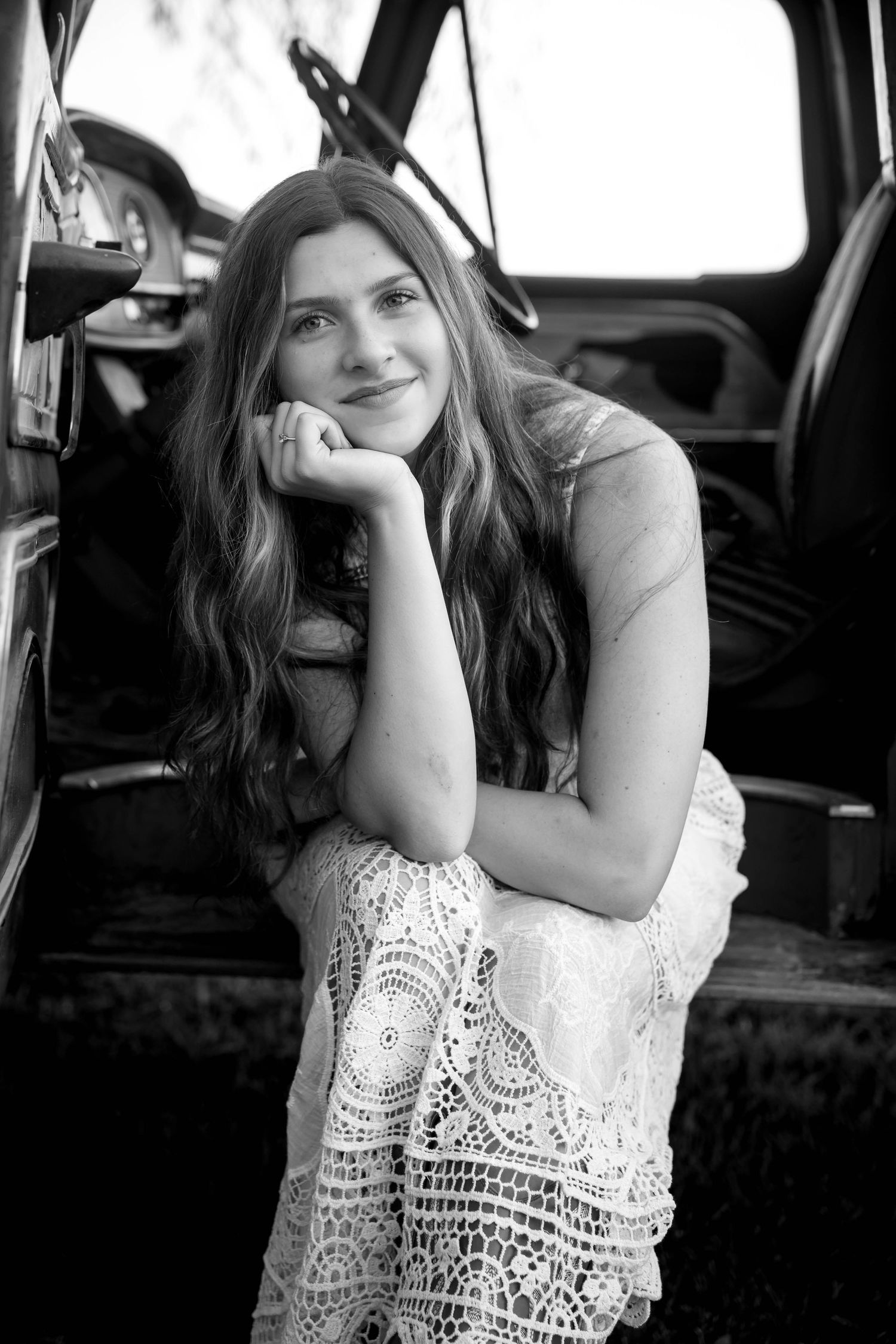 Young woman in lace dress smiling while sitting in vintage truck doorway, black and white portrait photo.