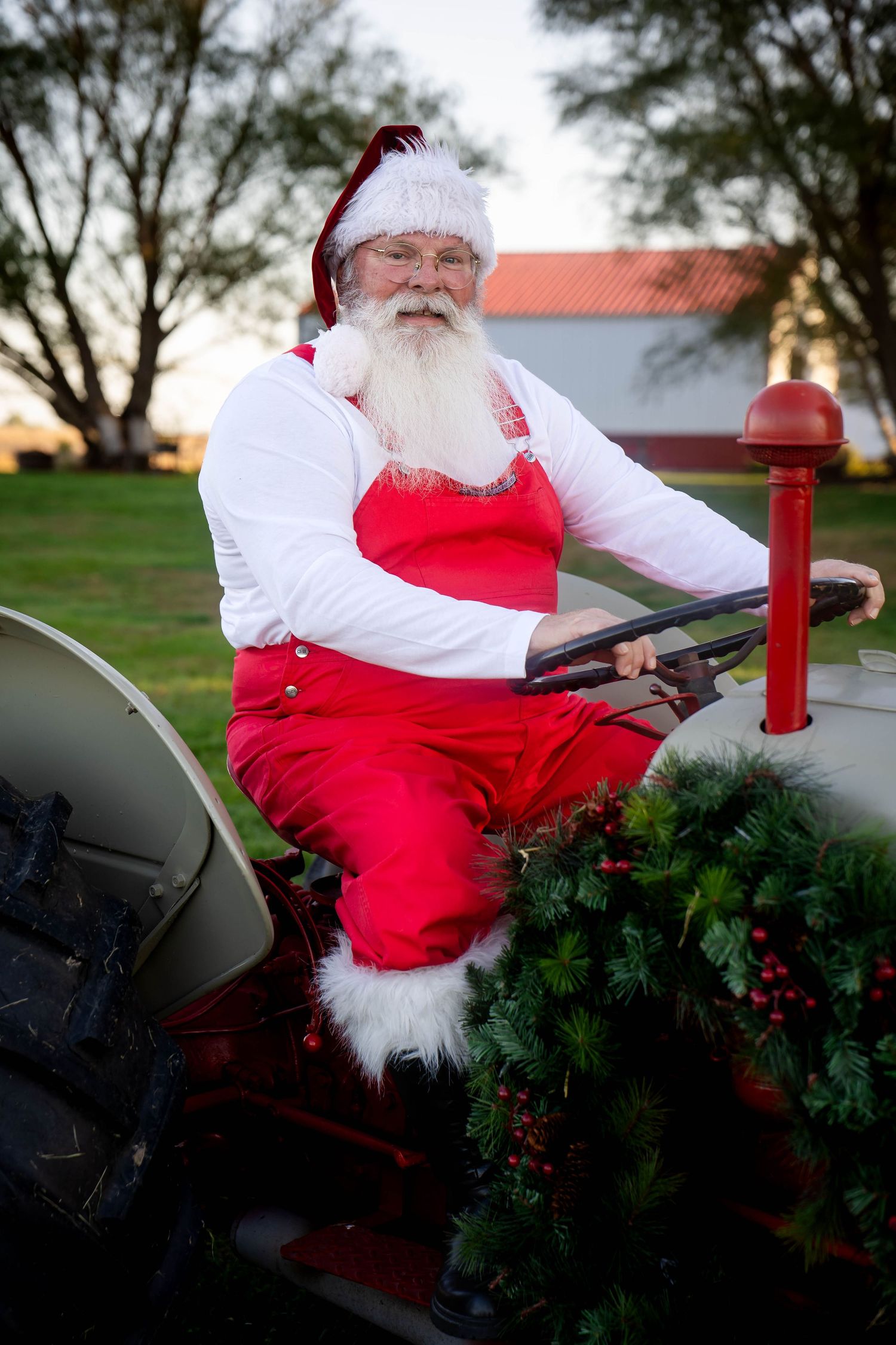 Santa Claus in red overalls sits on a decorated vintage tractor on a farm at sunset.