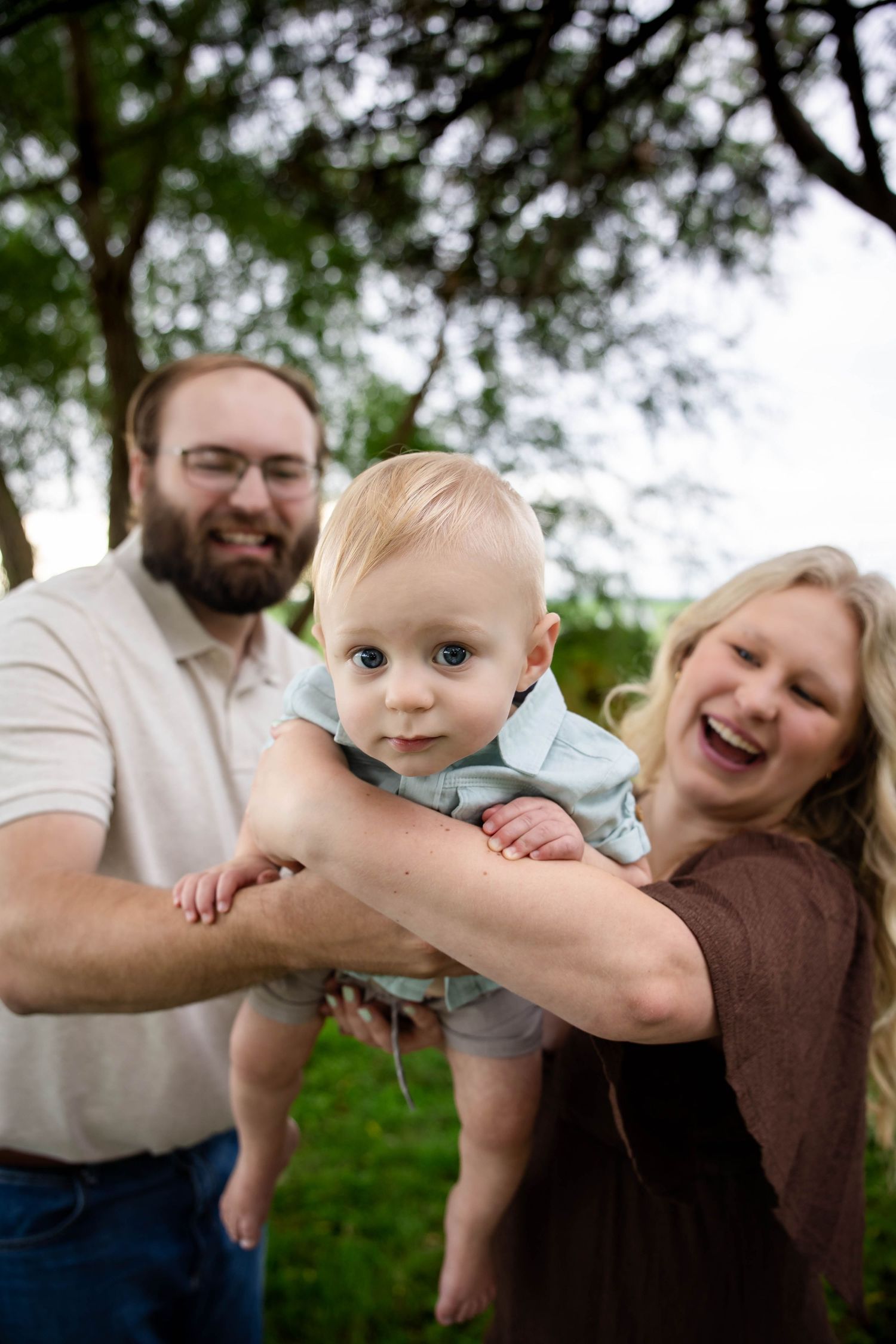 Happy parents lifting their blue-eyed baby outdoors under green trees in a joyful family portrait.