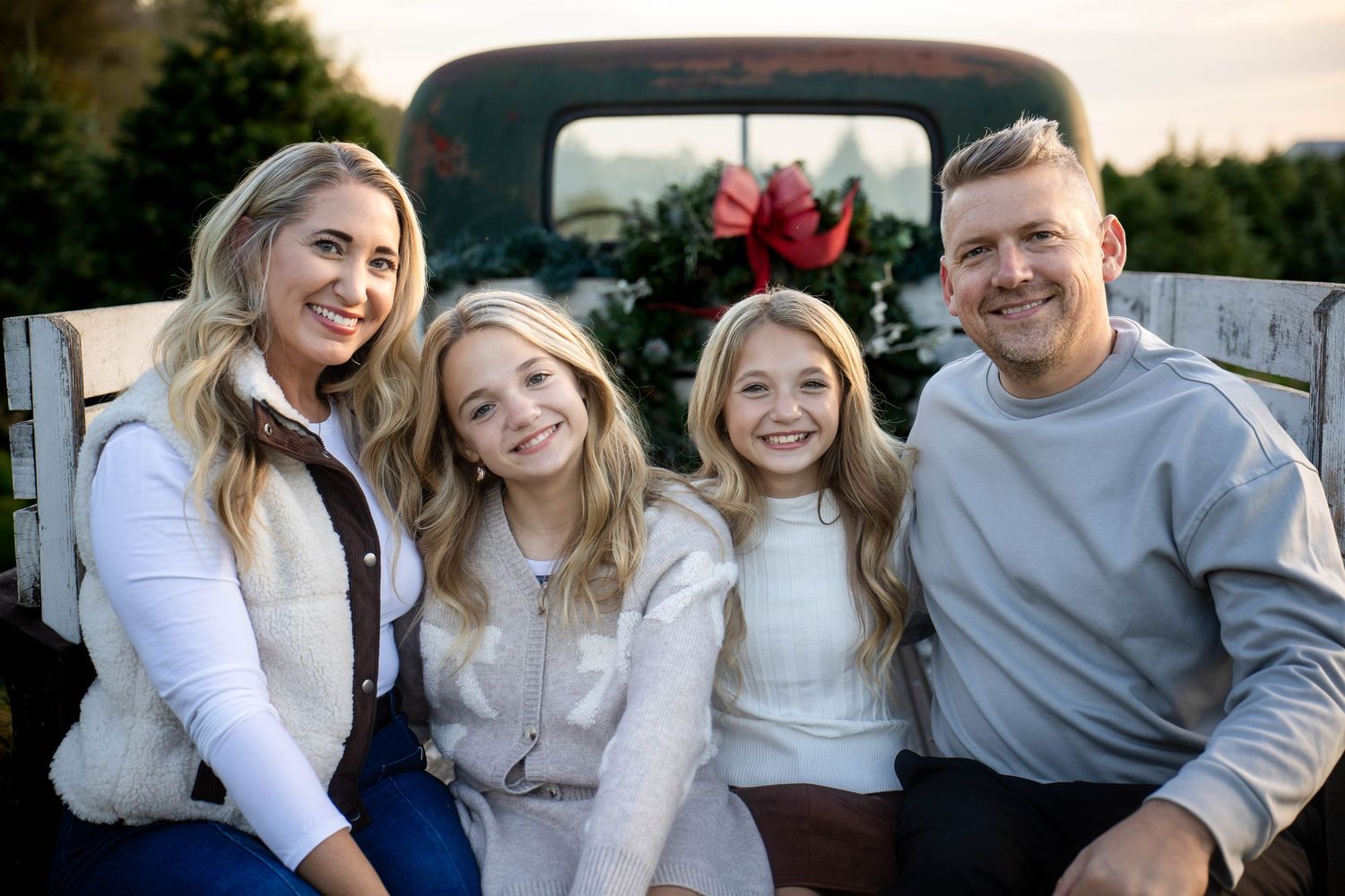 Smiling family of four posing together in front of a vintage truck decorated with a red bow and evergreen trees for a holiday photo.
