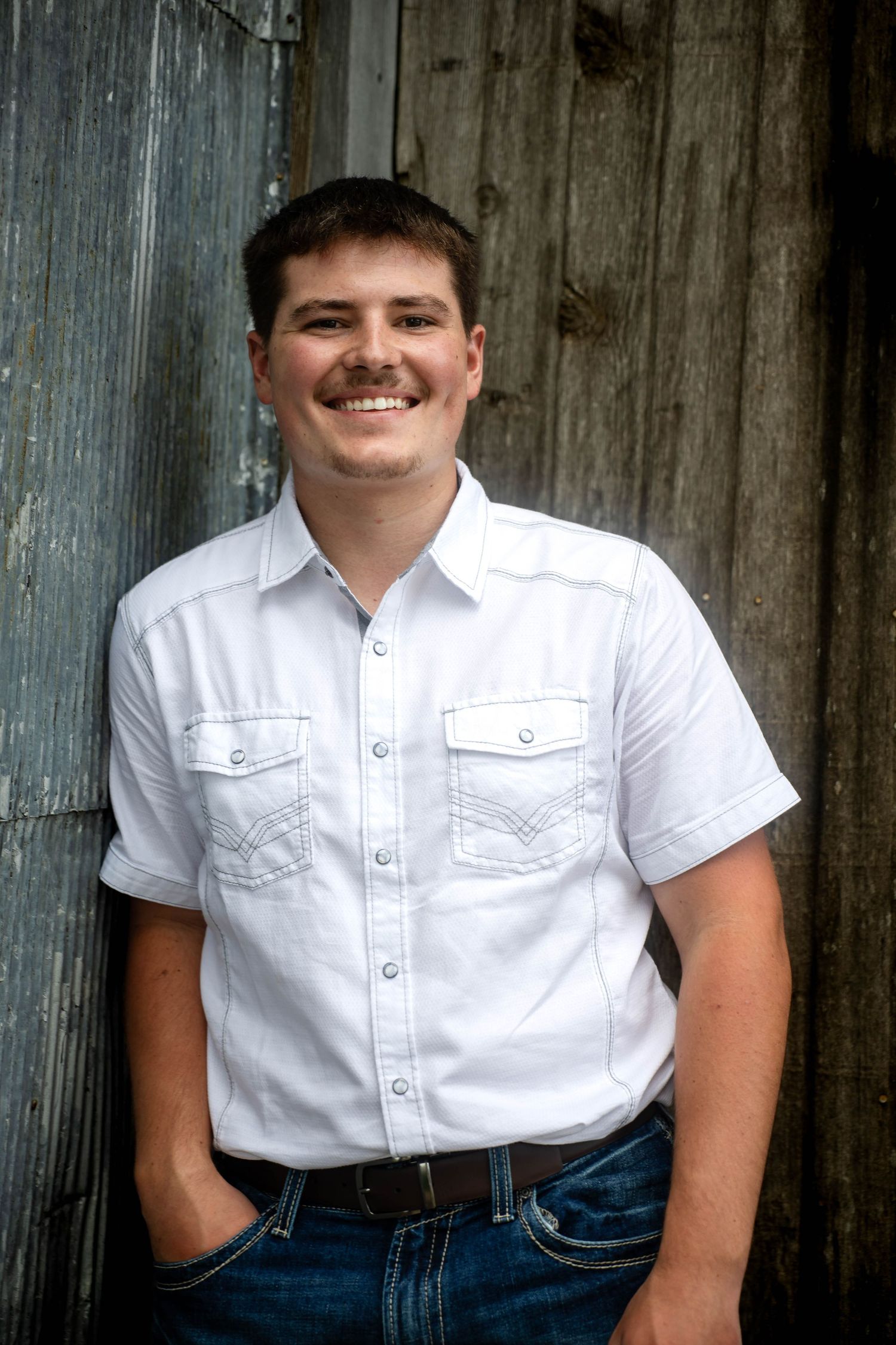 Smiling young man in a white button-up shirt and jeans leans against a weathered rustic wooden and corrugated metal wall.