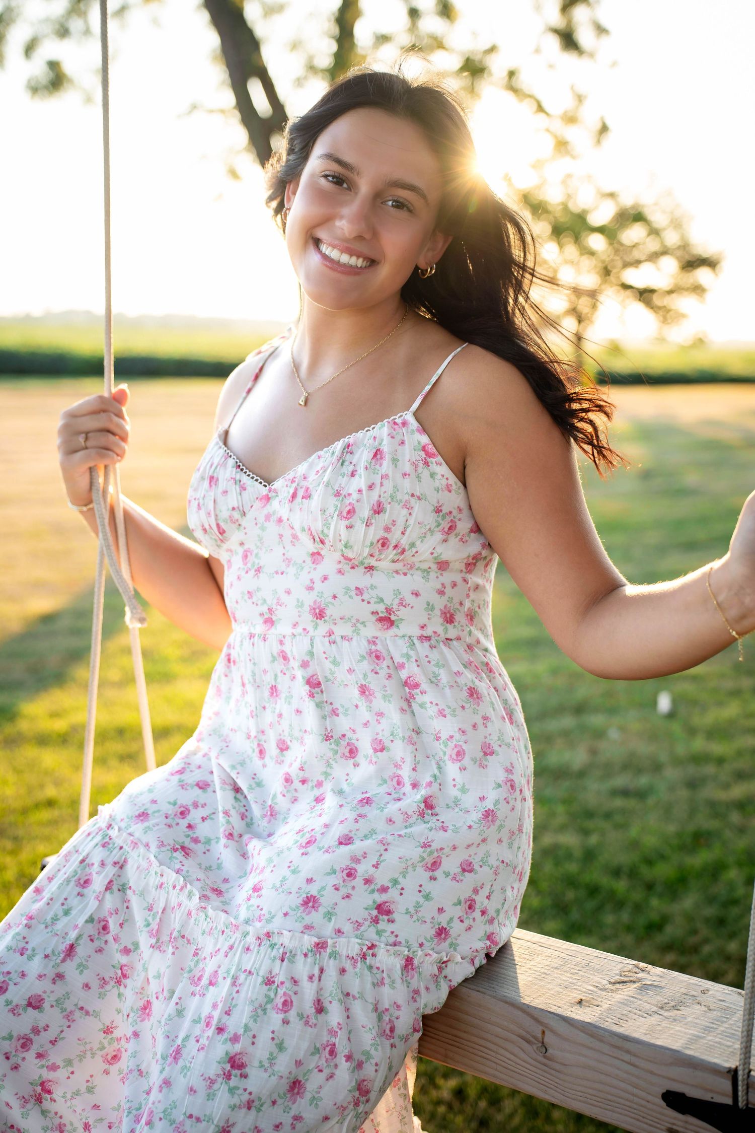 Smiling young woman in floral sundress sitting on a wooden swing at golden hour in a green field.