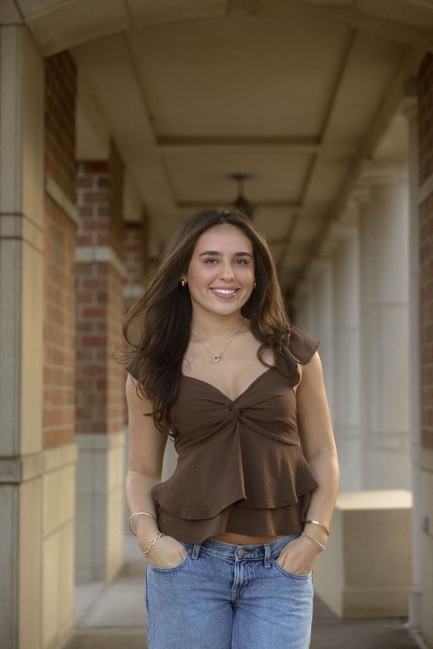 Smiling young woman with long dark hair in a brown ruffled top and light-wash jeans stands in a brick and stone covered walkway.