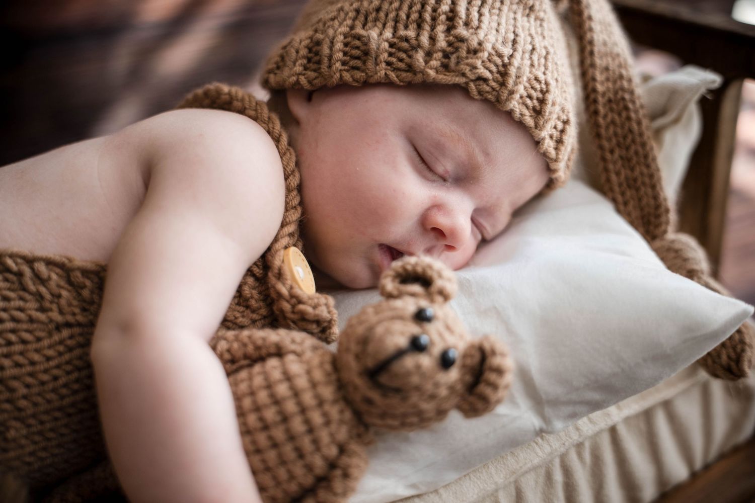 Sleeping newborn baby wearing a knit brown hat cuddles a small teddy bear in a cozy wooden crib.