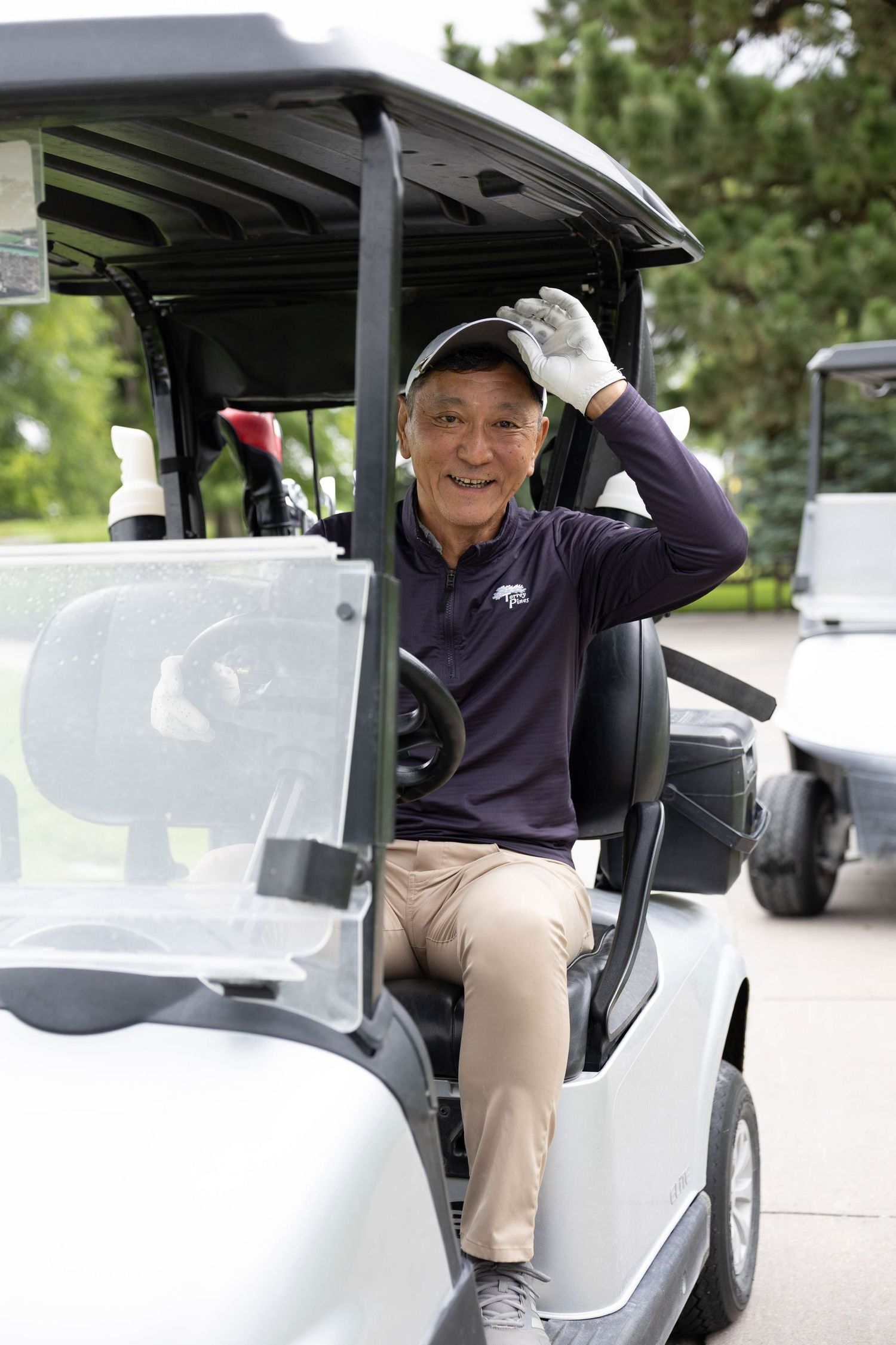 Smiling senior Asian man in golf attire waves from a white golf cart on a sunny course.
