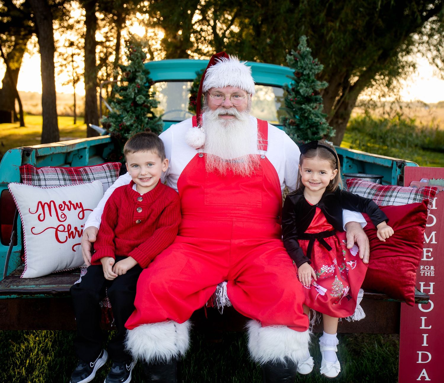 Santa Claus in a red suit sits on a vintage truck tailgate with a smiling boy and girl, with a Merry Christmas pillow and trees behind.