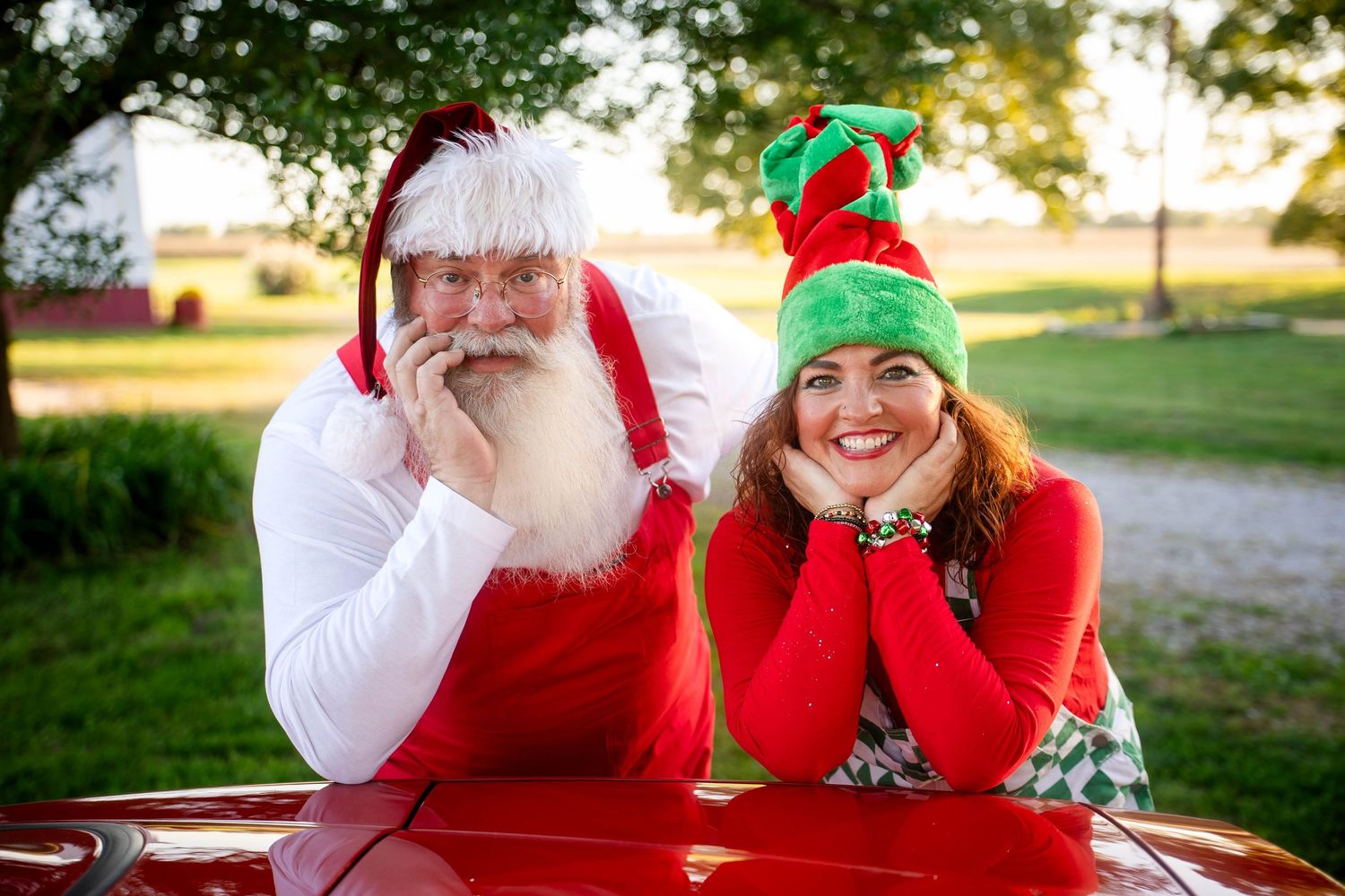 Santa Claus and a festive elf helper pose cheerfully together, leaning on a red vehicle outdoors.