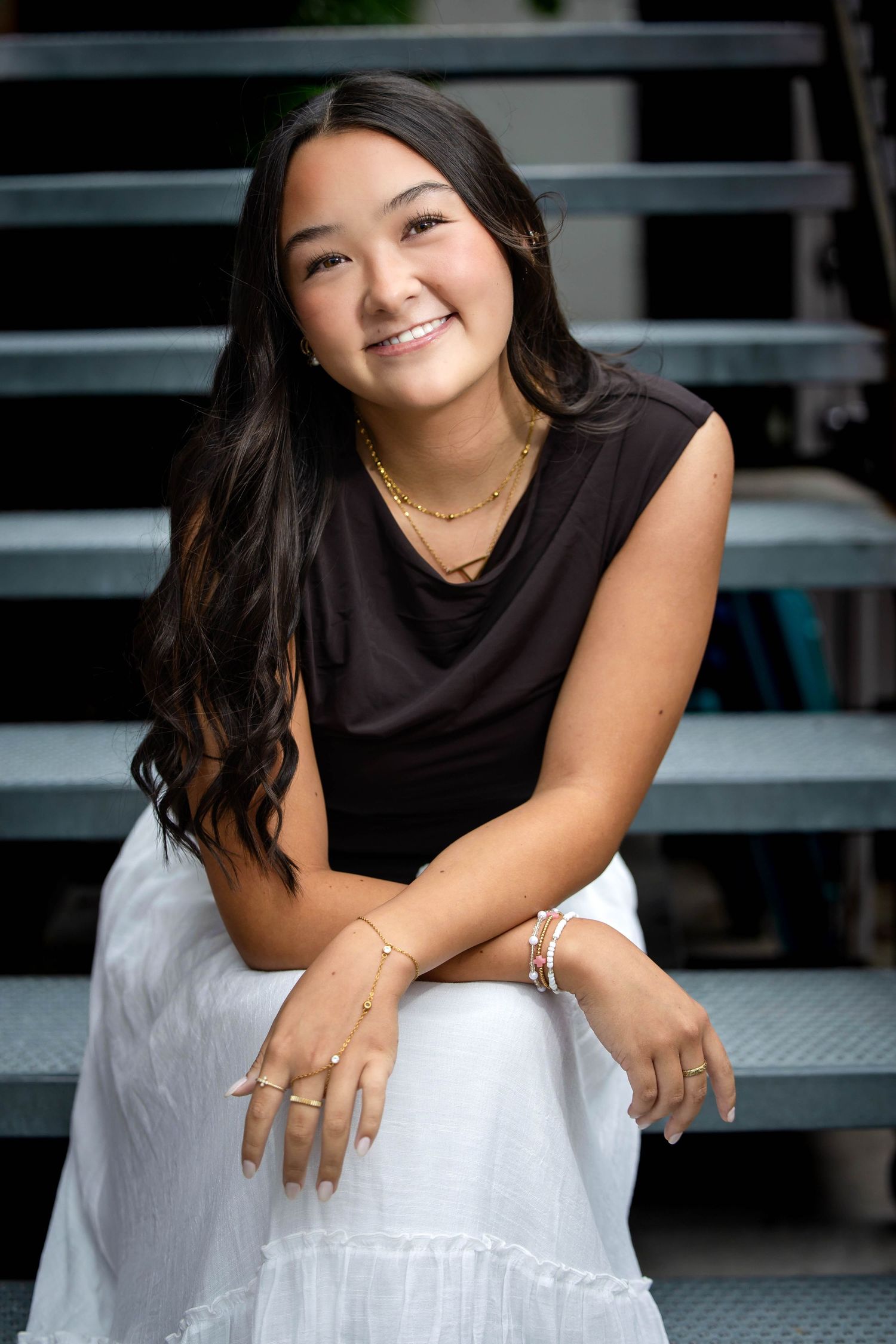 Smiling young woman with dark hair in black top and white skirt sitting on metal bleacher steps.
