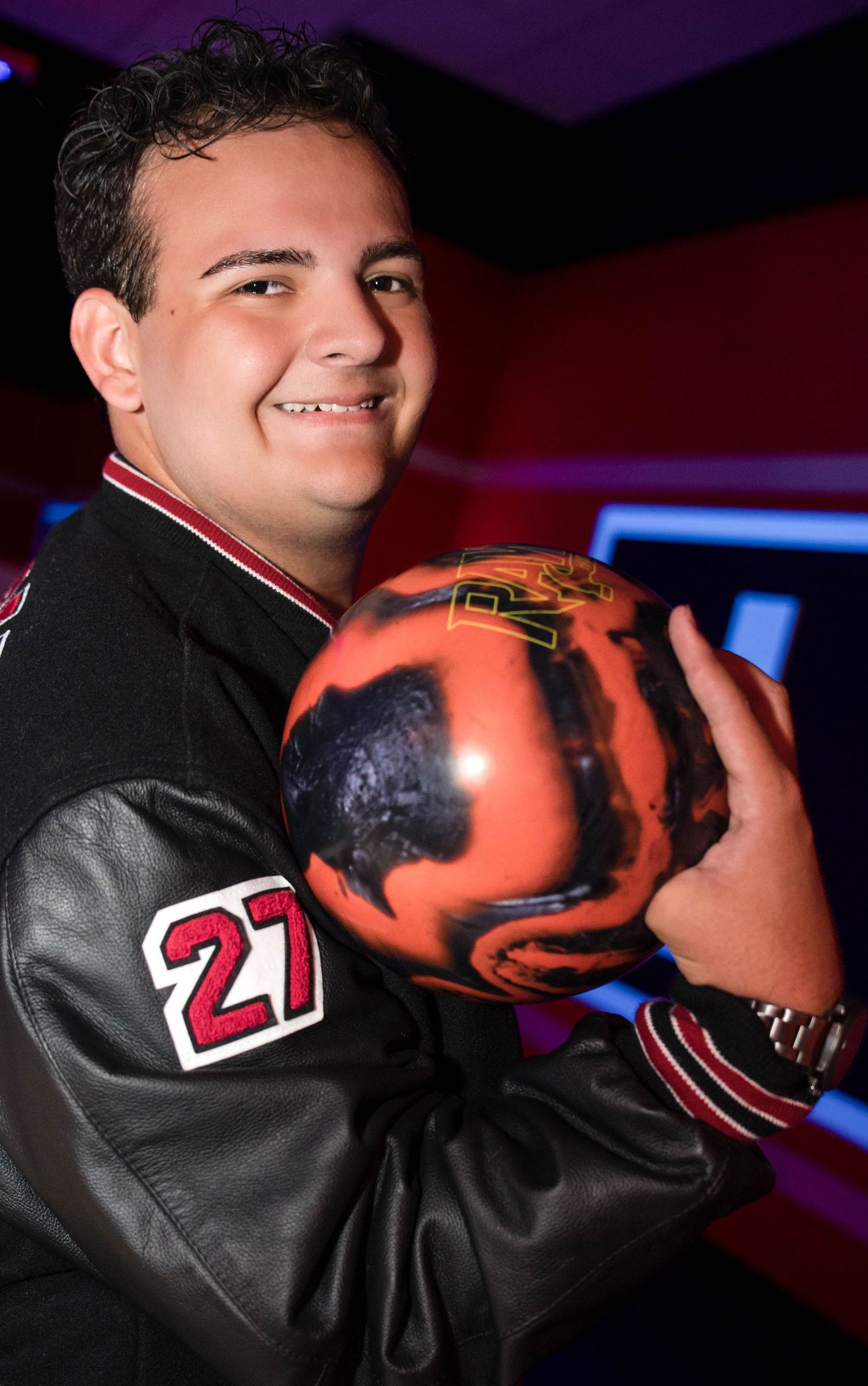 Teenage boy in letterman jacket number 27 holding orange bowling ball at bowling alley.
