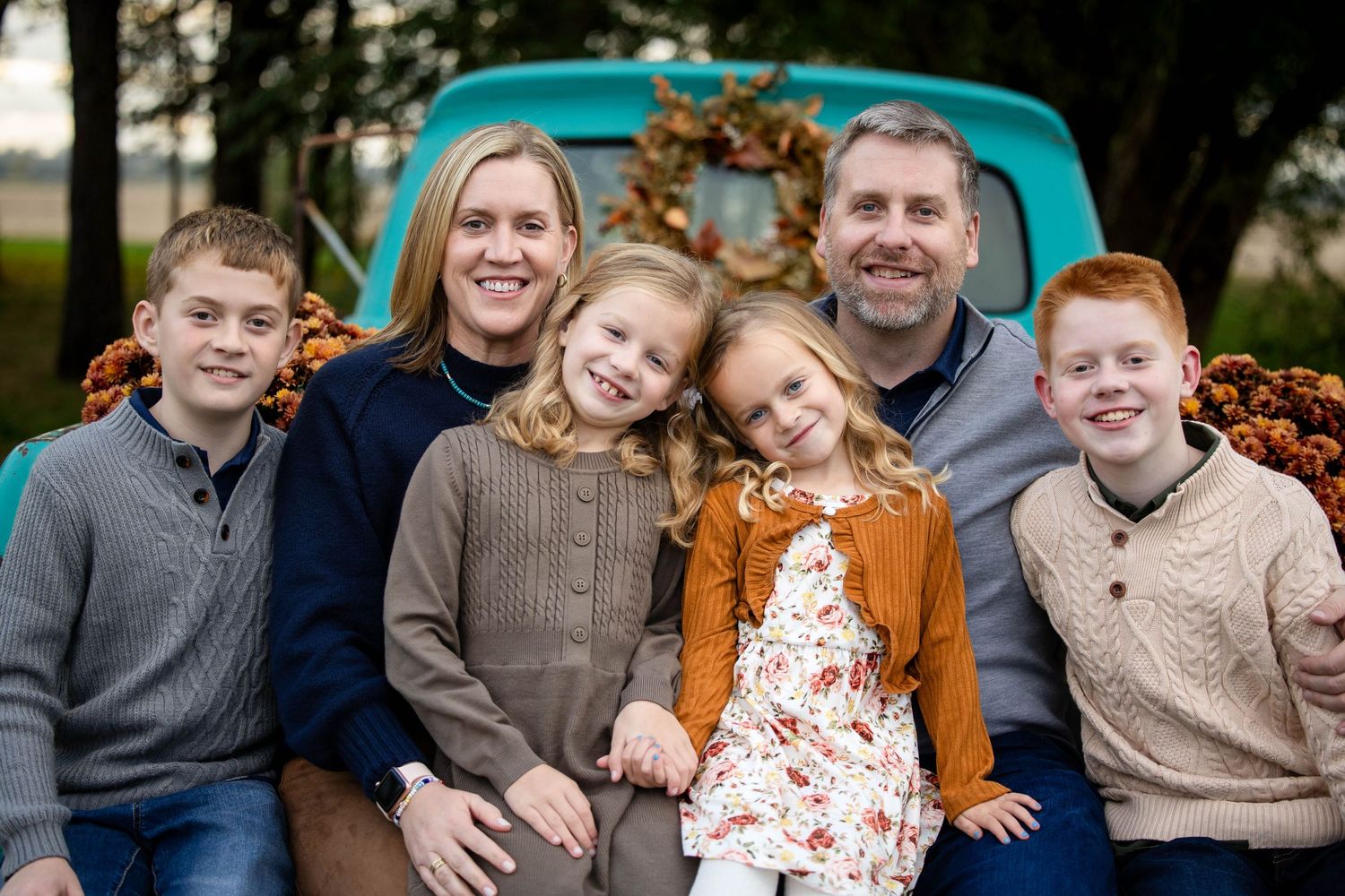 Smiling family of six in cozy fall sweaters poses together in front of a vintage teal truck decorated with a wreath and orange pumpkins.