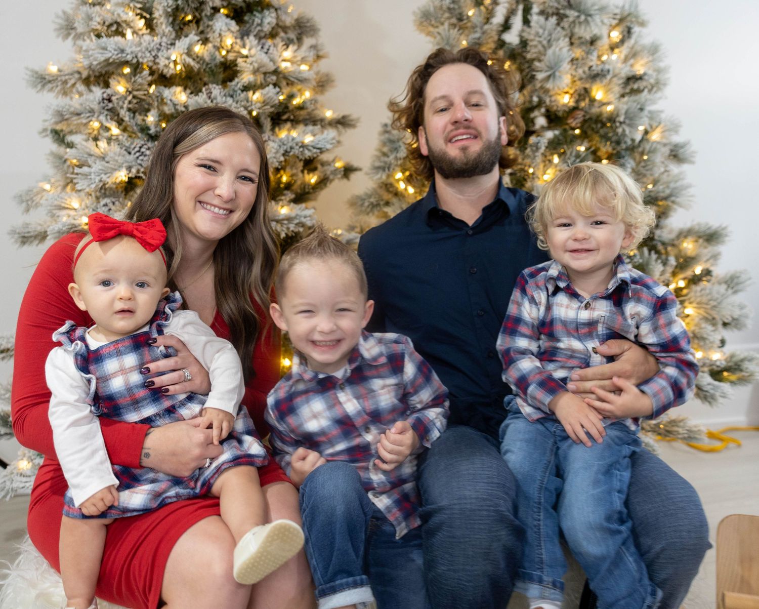 Smiling family of five in matching plaid outfits posing together in front of decorated, softly lit Christmas trees for a holiday portrait.