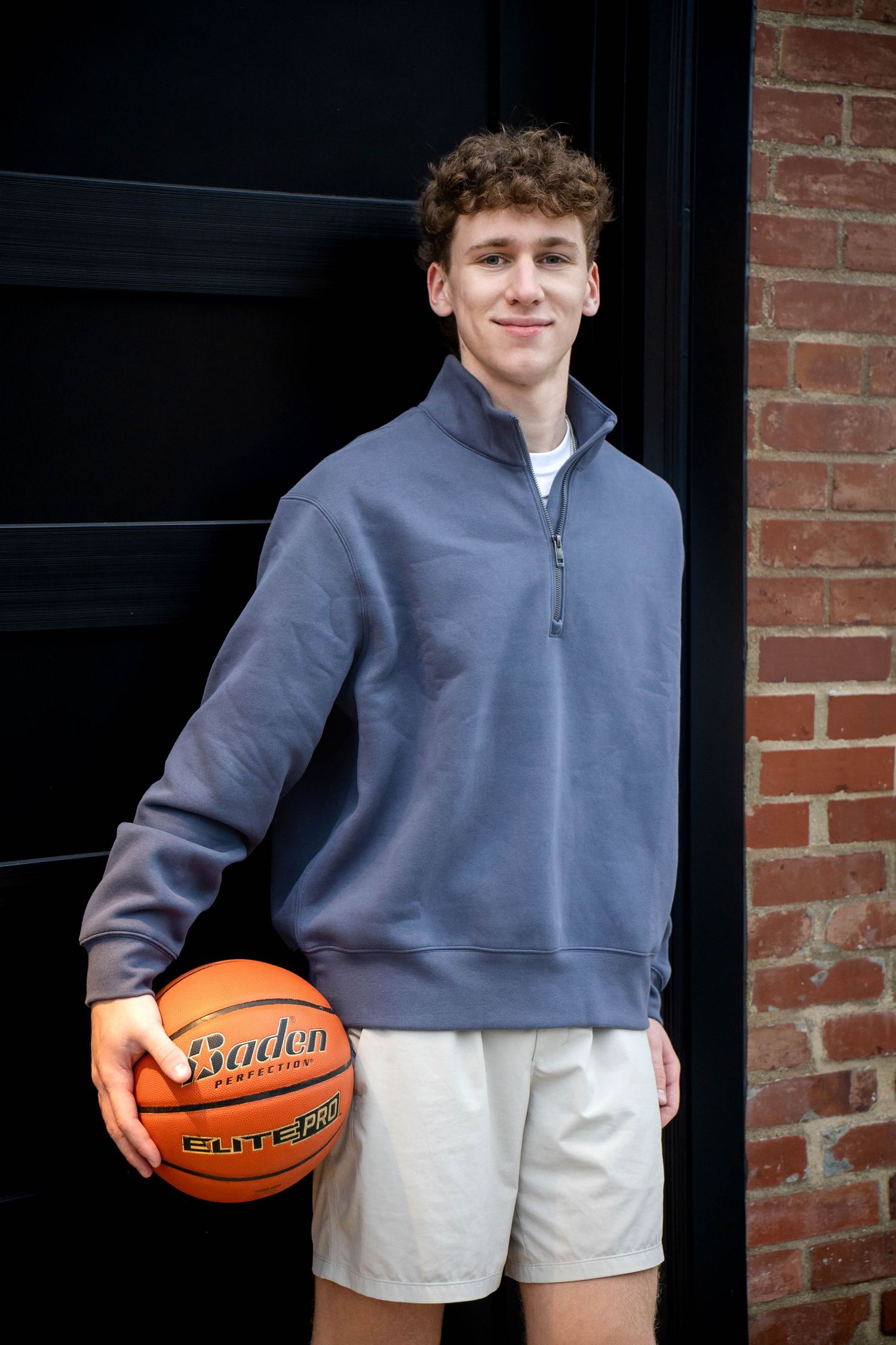 Smiling young man with curly hair in a gray quarter-zip pullover holds a Baden basketball beside a brick wall.