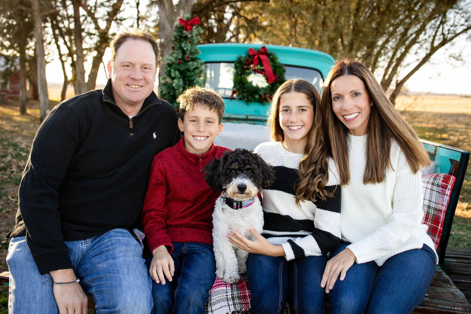 Family of four smiling with their dog in the bed of a vintage teal truck decorated for Christmas.