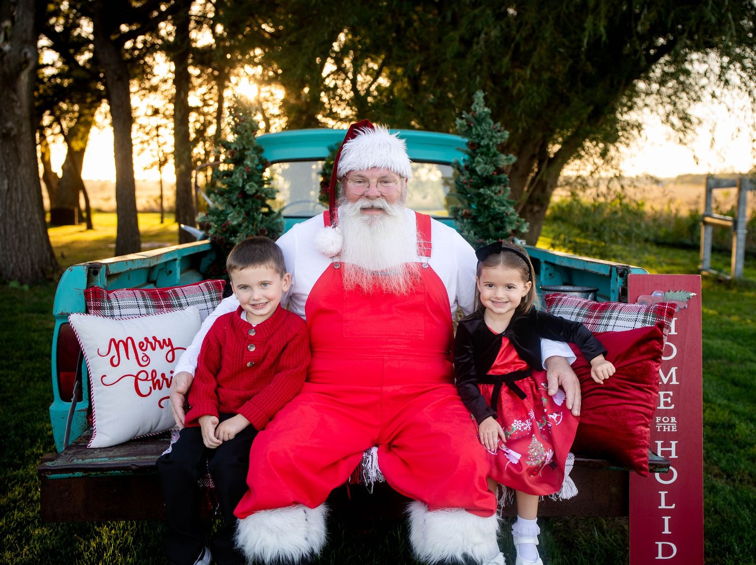 Two children pose with Santa Claus in a vintage teal truck bed decorated for Christmas outdoors.