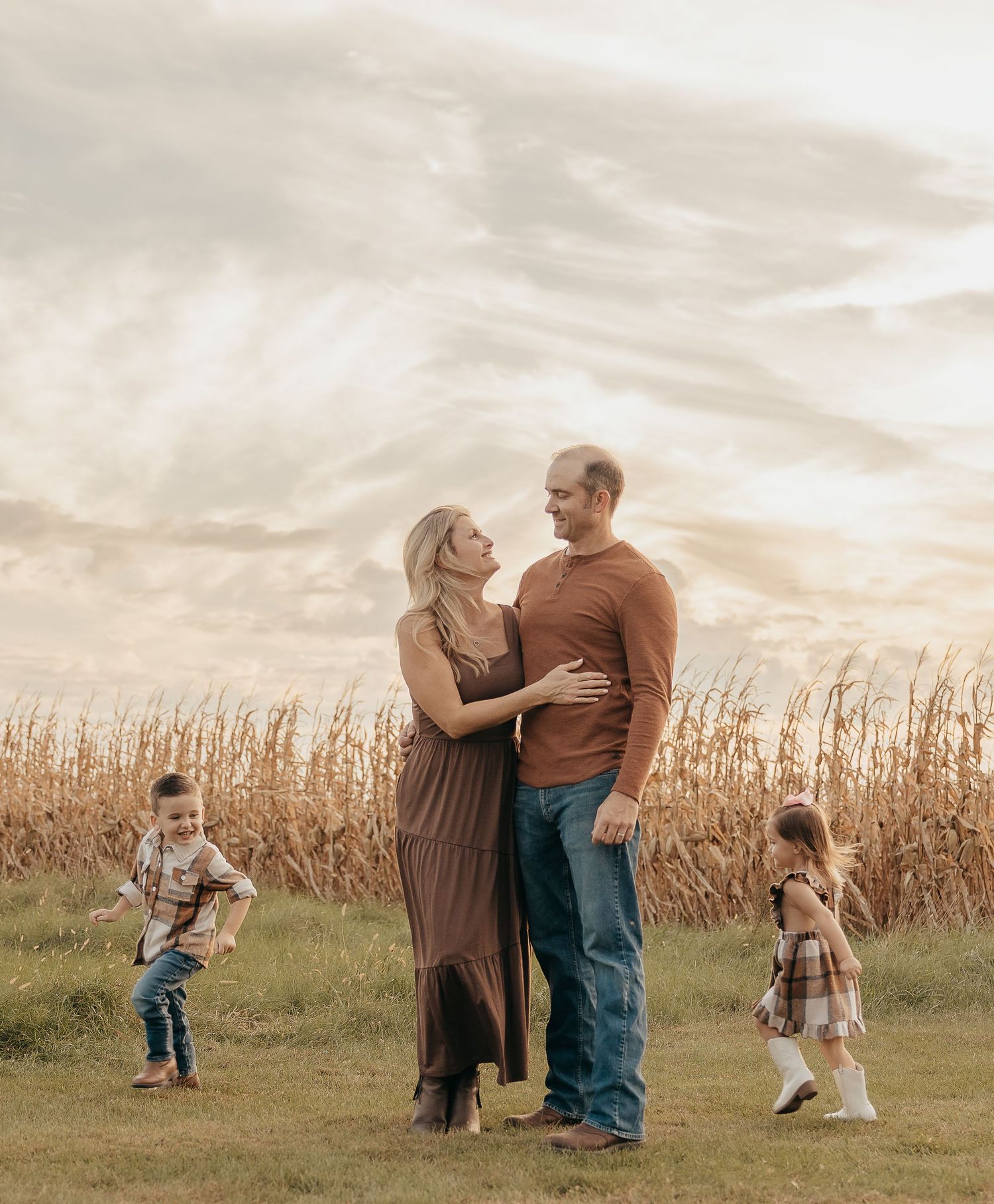 Family portrait with couple embracing and two young children playing in field near golden cornfield at dusk.