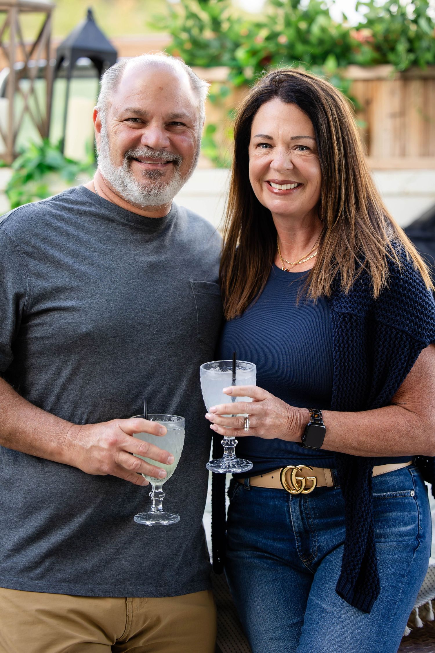 Smiling middle-aged couple holding wine glasses pose together at an outdoor garden party.
