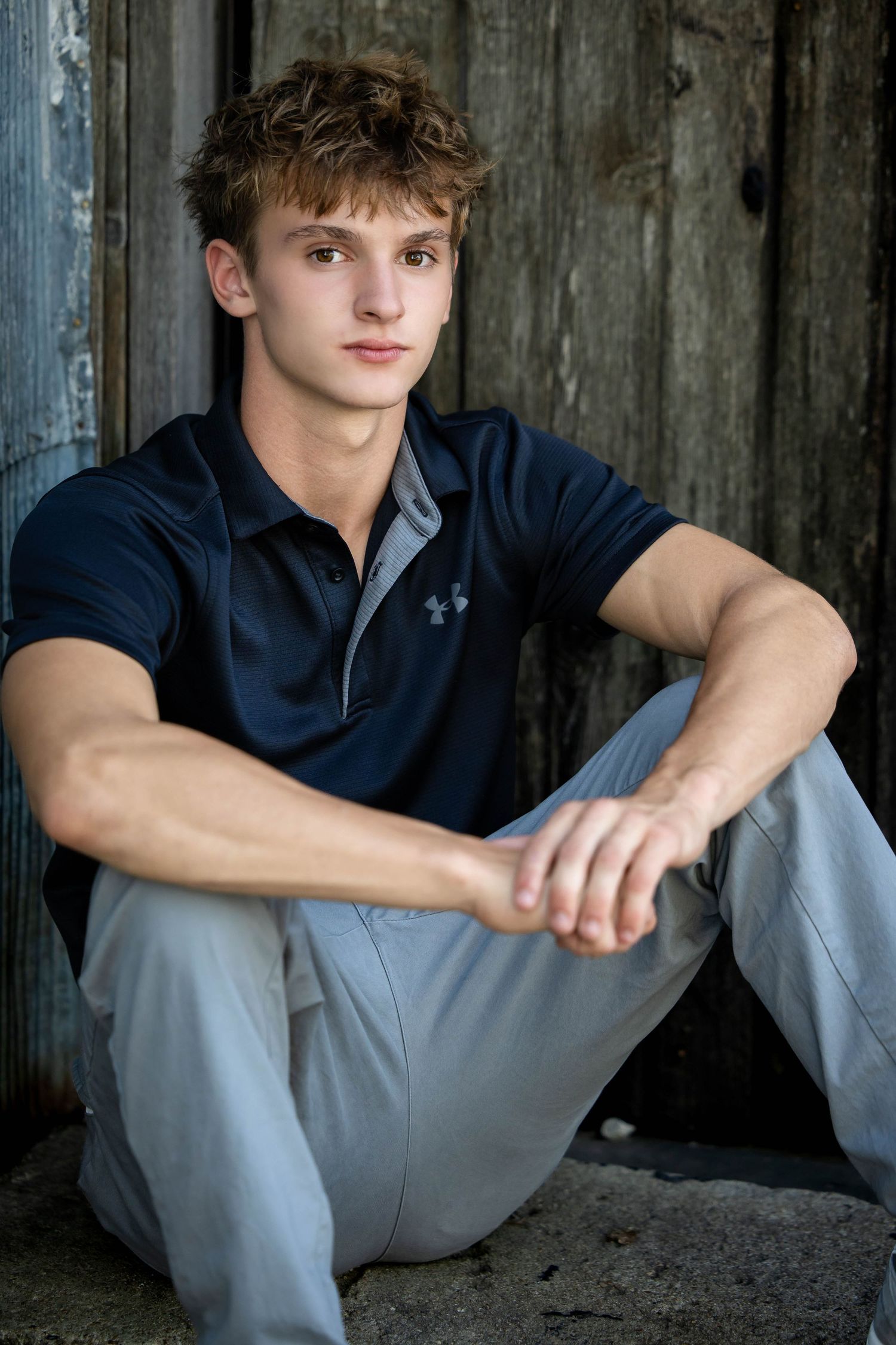 Young man in a navy polo and gray pants sits on a concrete ledge with his arms resting on his knees in front of a weathered wooden wall.