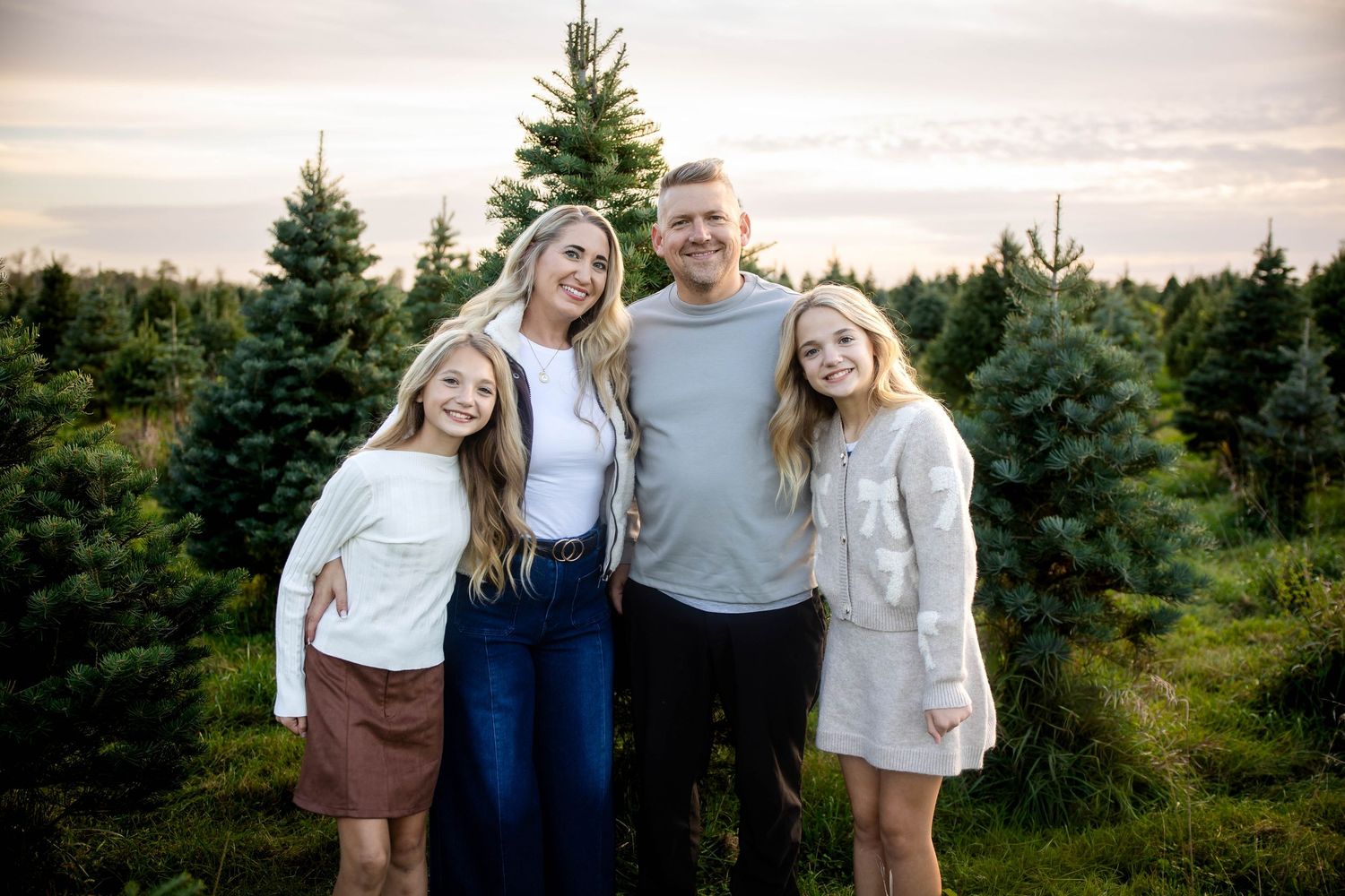 Smiling family of four, two adults and two girls, posing together in a Christmas tree farm at sunset.