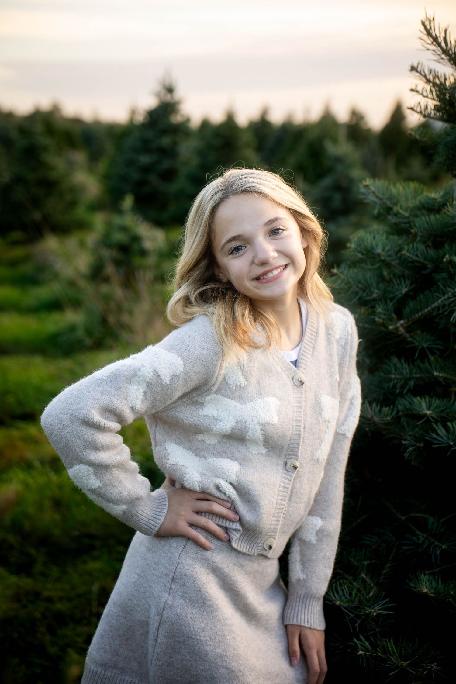 Smiling girl with long blonde hair in a cozy gray cloud sweater and skirt poses with hand on hip beside evergreen trees at sunset.