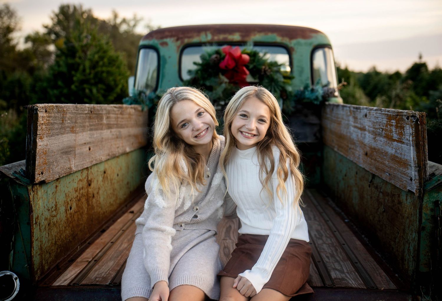 Two smiling girls with long blonde hair sit together on the wooden bed of a vintage green truck decorated with a red-bowed holiday wreath.