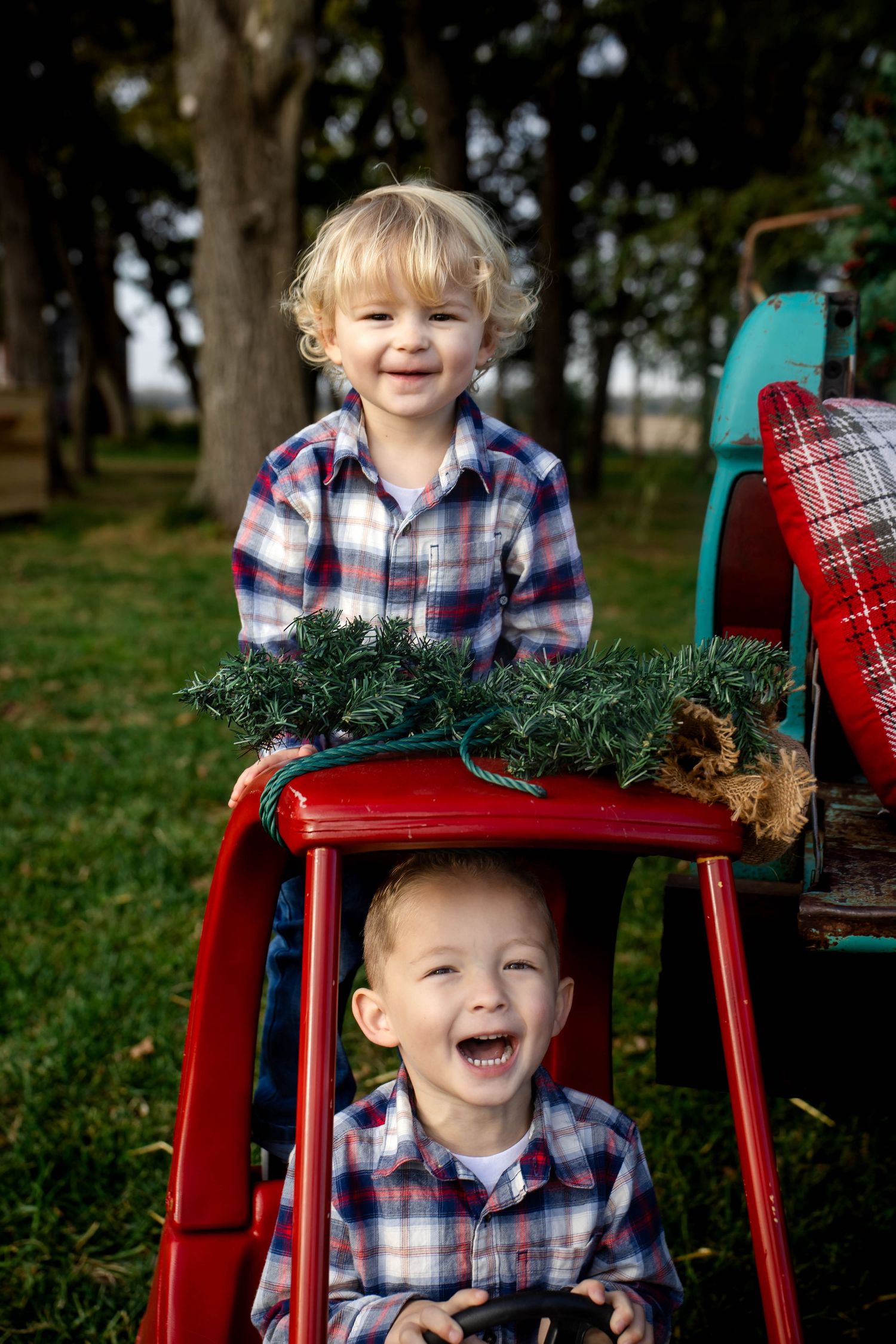 Two laughing toddler boys in plaid shirts play with a red vintage toy truck decorated with a pine garland in a sunny outdoor setting.