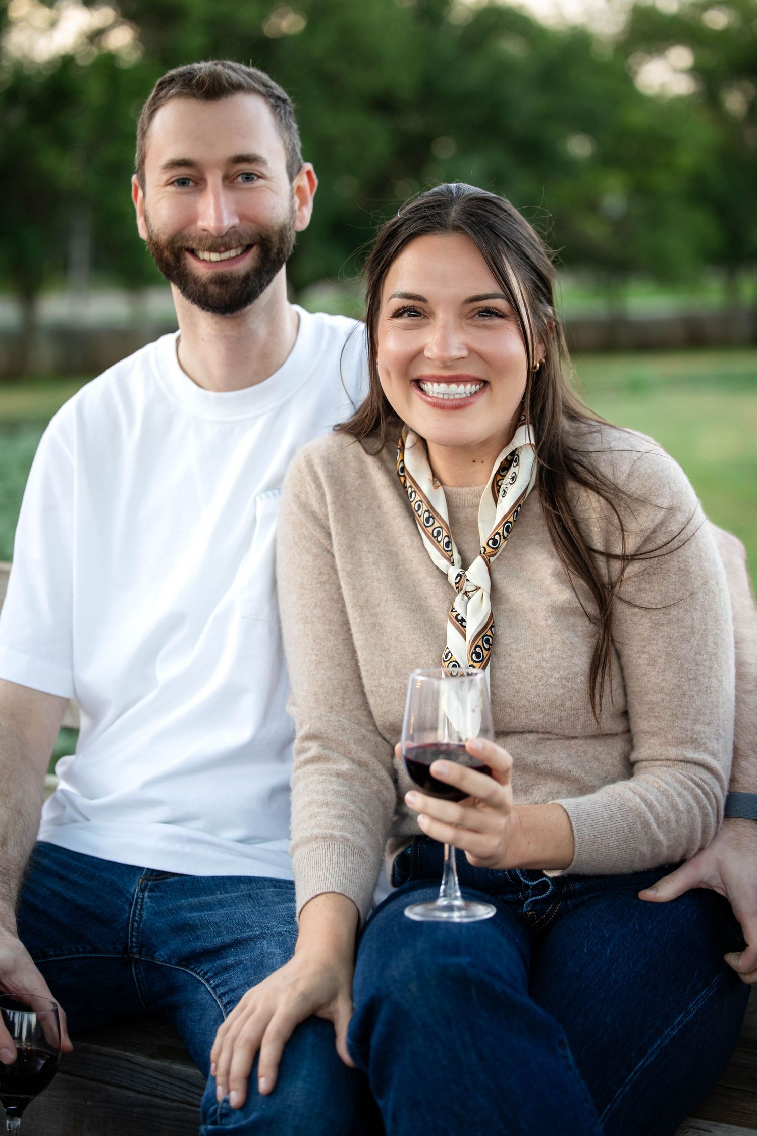 Smiling couple enjoying wine outdoors in a lush green park setting, woman holding a glass of red wine.