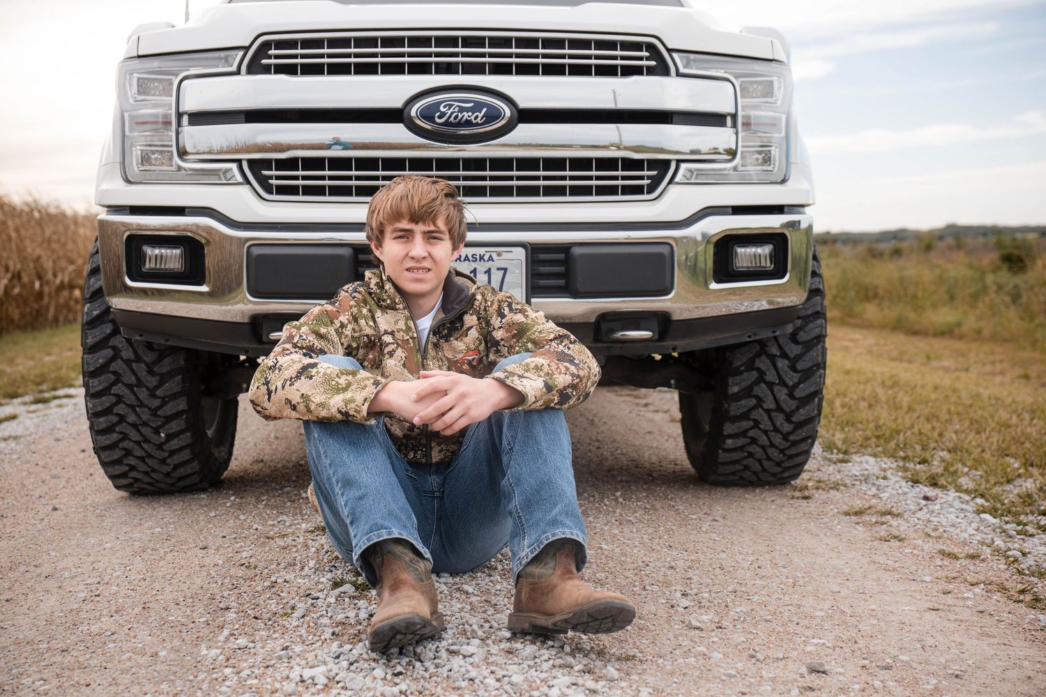 Young man in camouflage jacket and jeans sits on a gravel road in front of a white Ford truck with a grassy field behind.