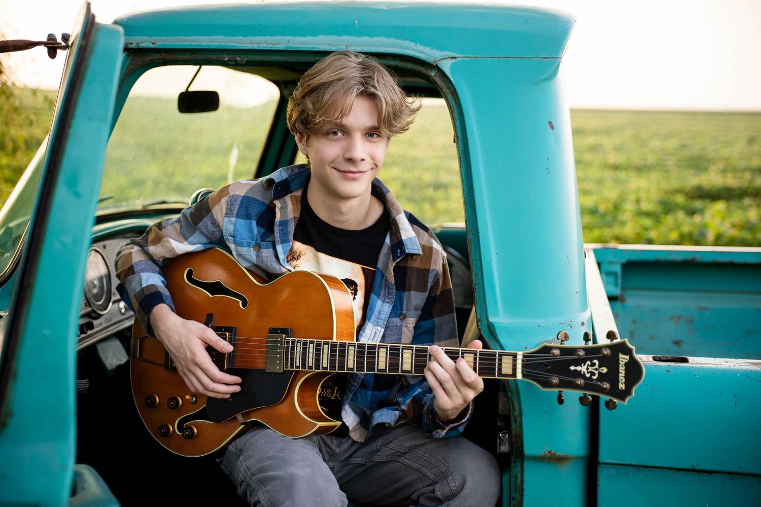 Teen boy playing guitar while sitting in a vintage teal pickup truck in a rural farm field.