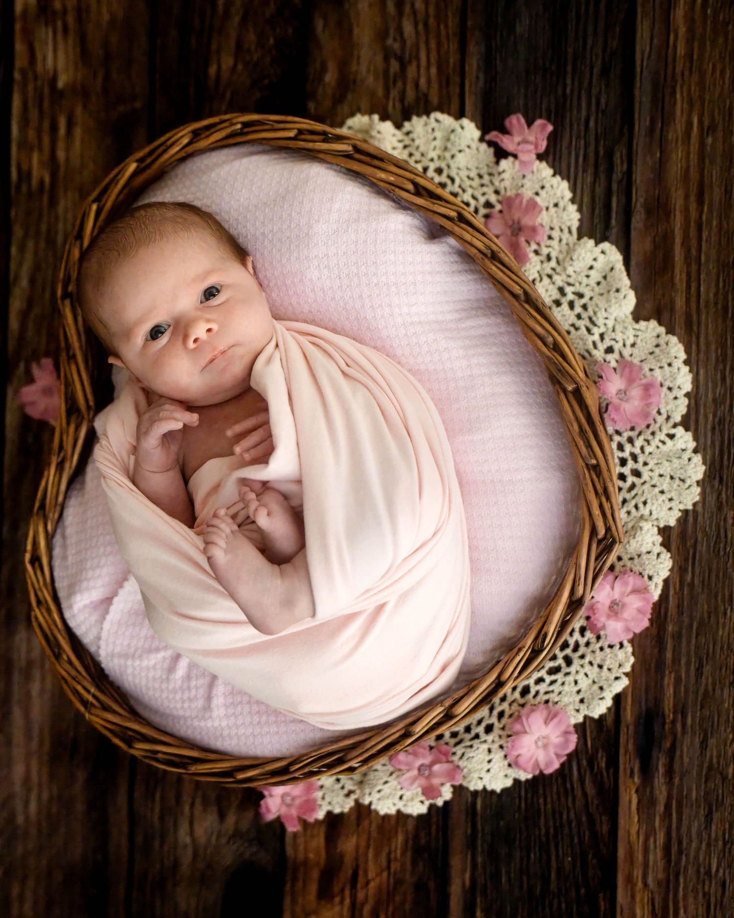 Newborn baby wrapped in a soft pink swaddle lying in a wicker basket with lace trim and pink flowers on a rustic wooden floor.