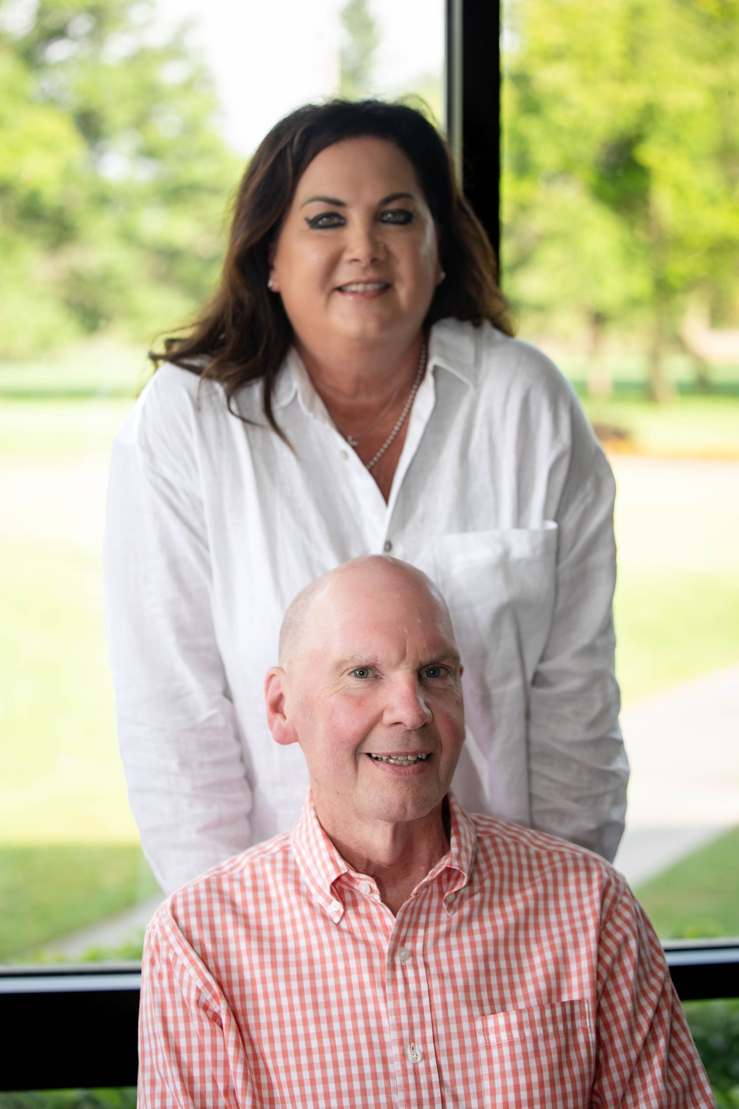 A smiling woman in white stands behind a seated older man in a pink plaid shirt near a bright window.