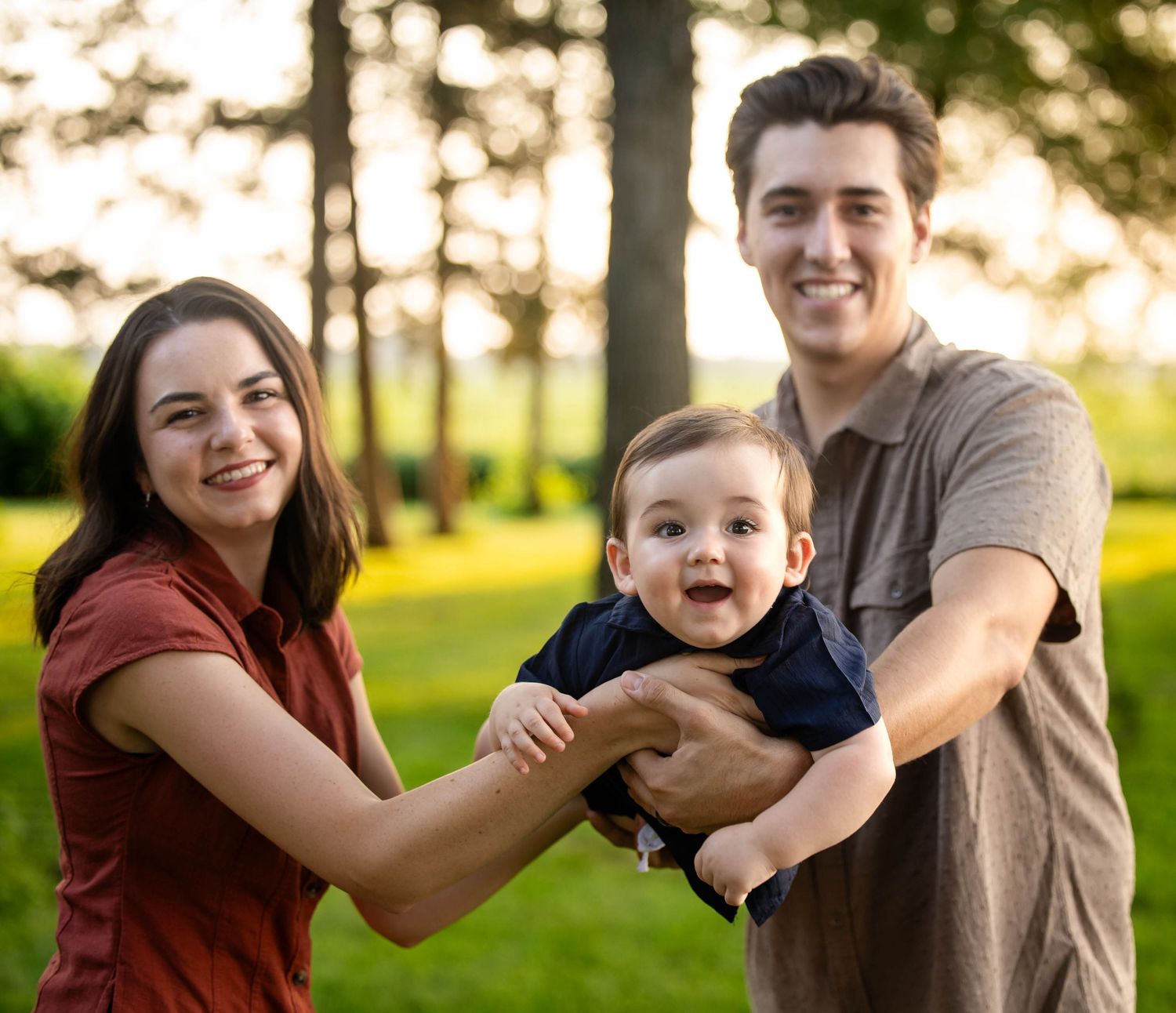 Young couple holding a laughing baby boy outdoors in a green park with warm sunset lighting.
