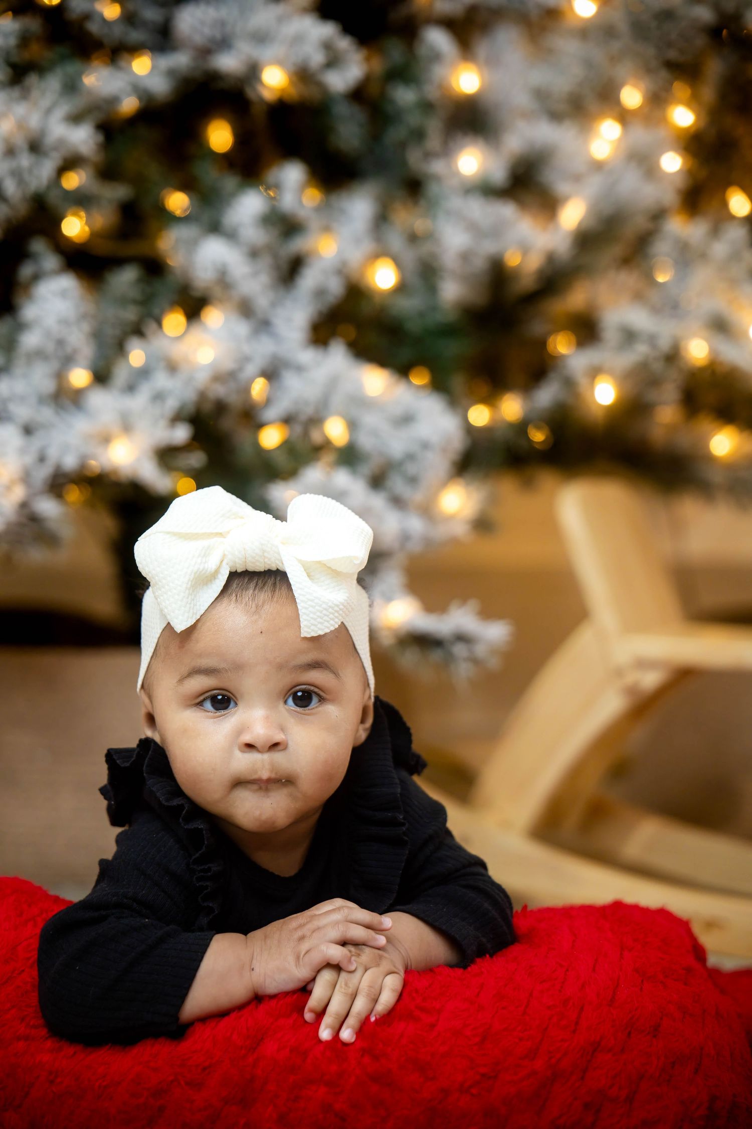 Baby in a black outfit and cream bow headband lies on a red blanket in front of a frosted Christmas tree with warm glowing lights.