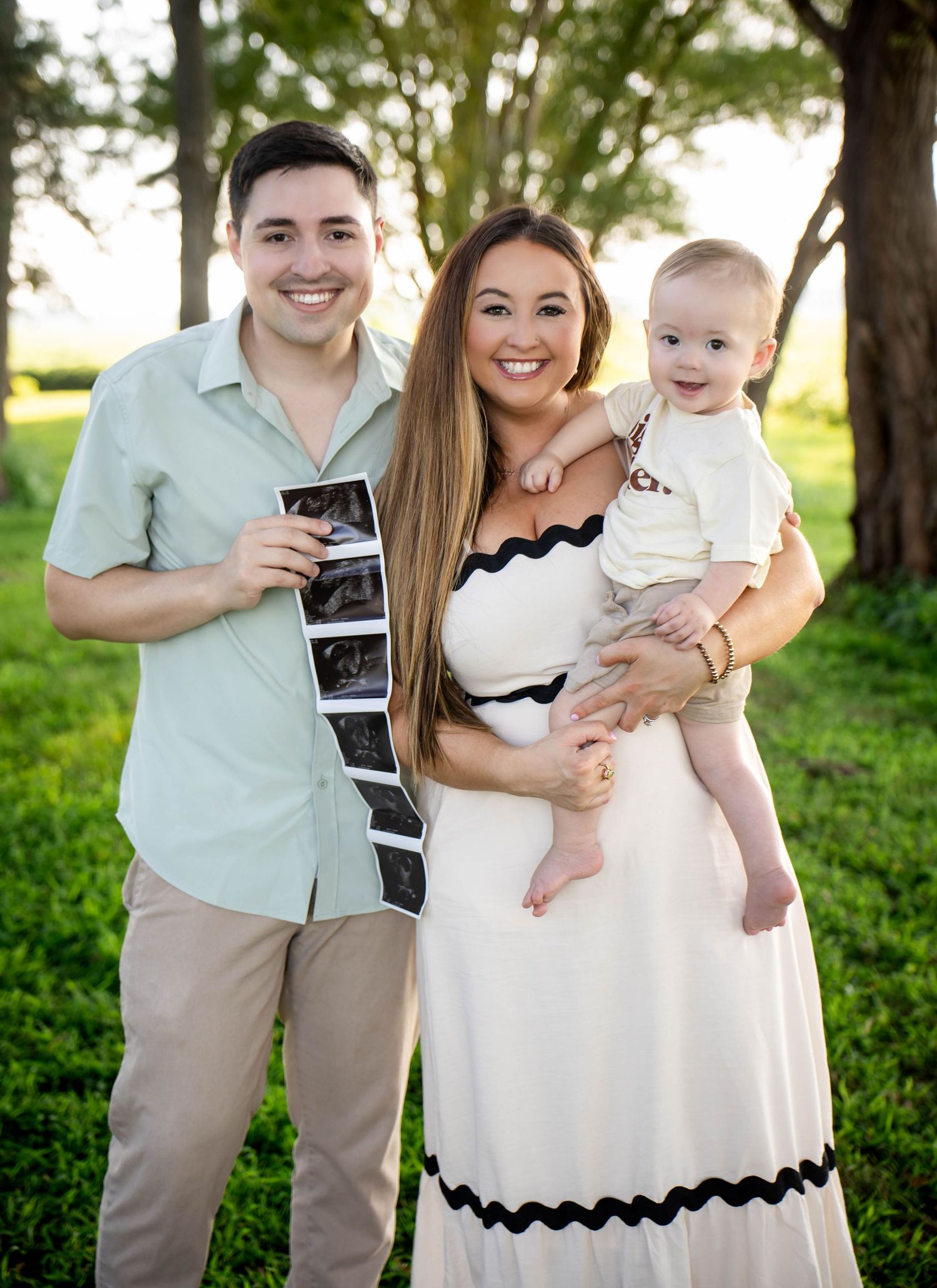 Smiling couple holding ultrasound photos with their baby outdoors for a pregnancy announcement.