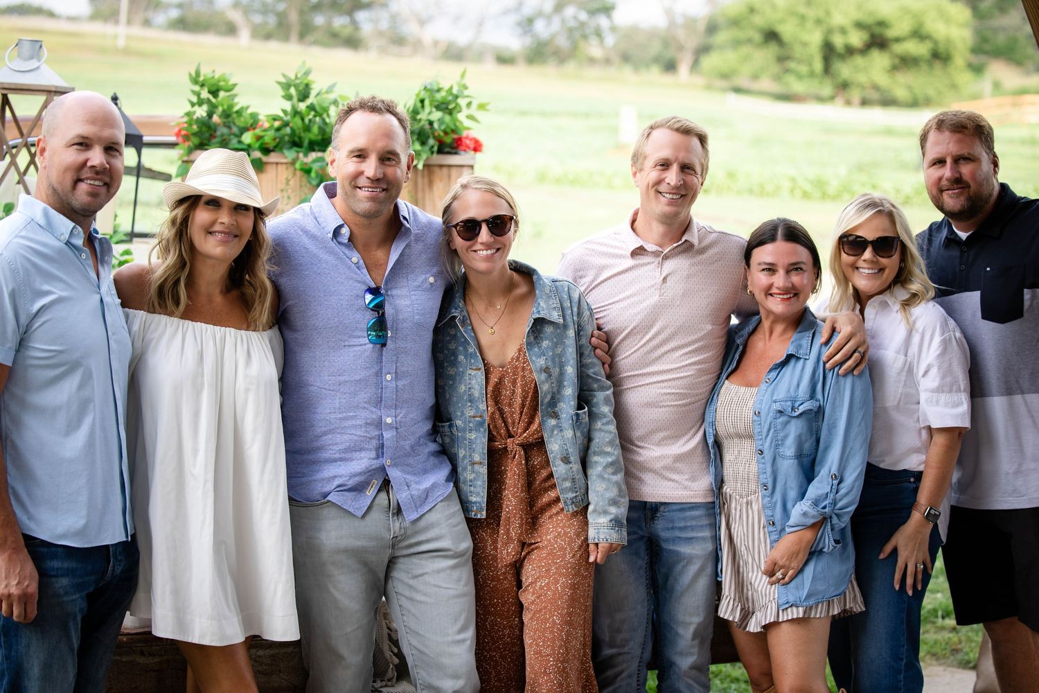 Group of eight smiling adults pose together for a photo at a sunny outdoor social gathering.